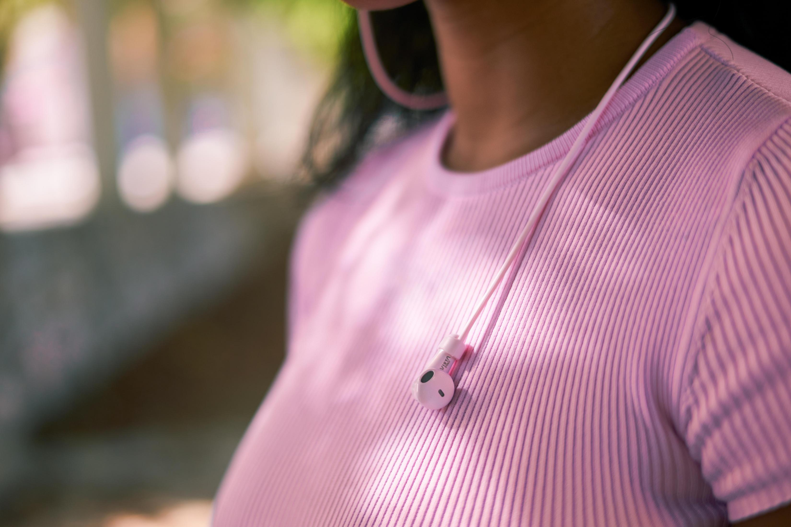 Person wearing pink shirt with pink earbuds, white cable. Blurred background.