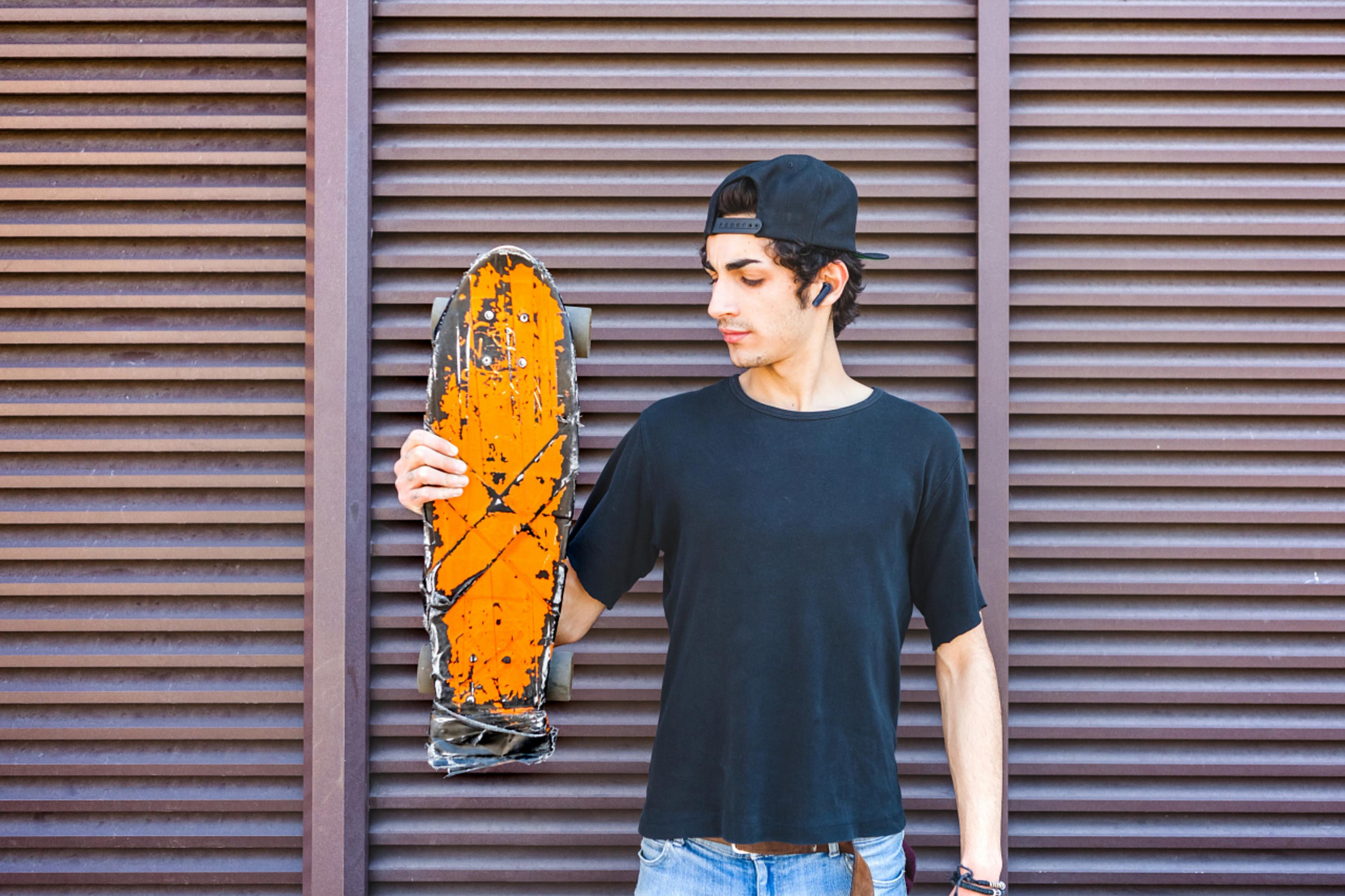Mężczyzna ze deskorolką przed brązową żaluzją. Nosi czapkę, koszulę i dżinsy.