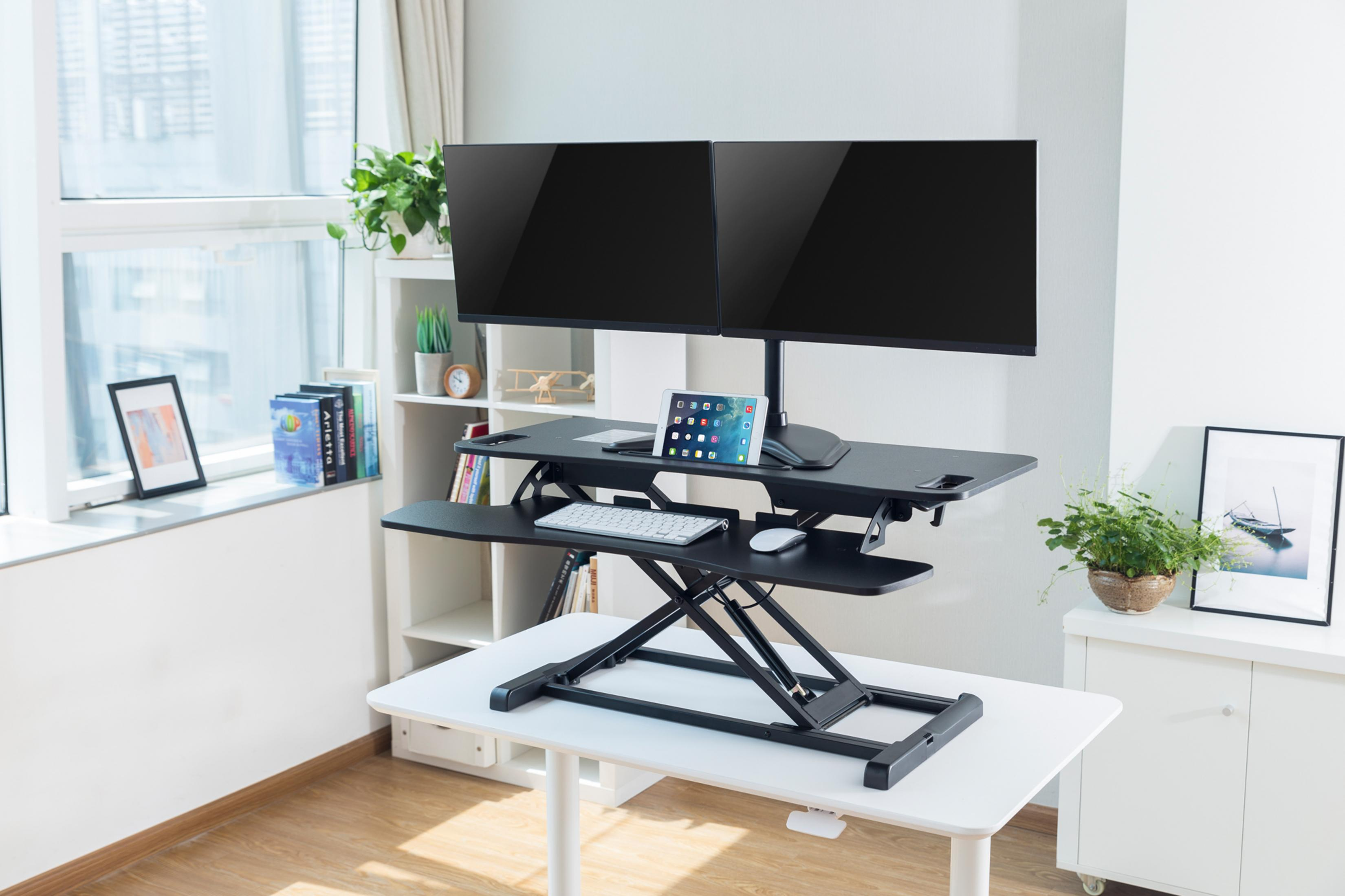 Biurko z regulacją wysokości z dwoma monitorami, tabletem, klawiaturą, roślinami i oknem.