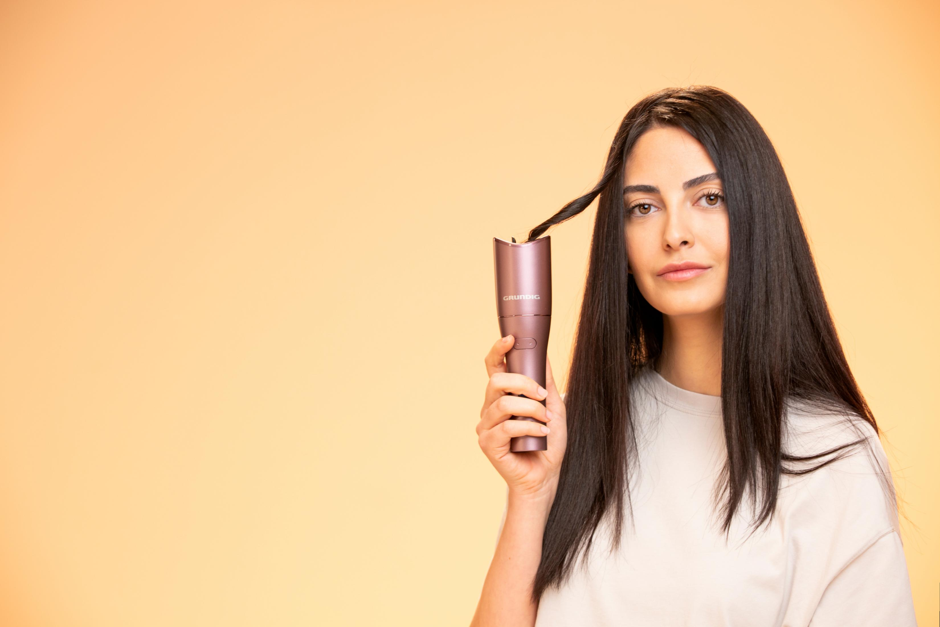 Donna con lunghi capelli scuri tiene un arricciacapelli, guardando la fotocamera con espressione neutra.