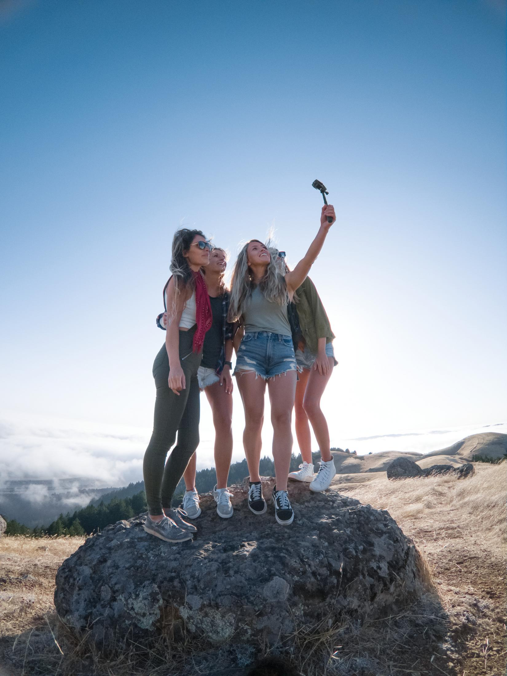 Vier Frauen machen ein Selfie auf einem Felsen, blauer Himmel im Hintergrund.