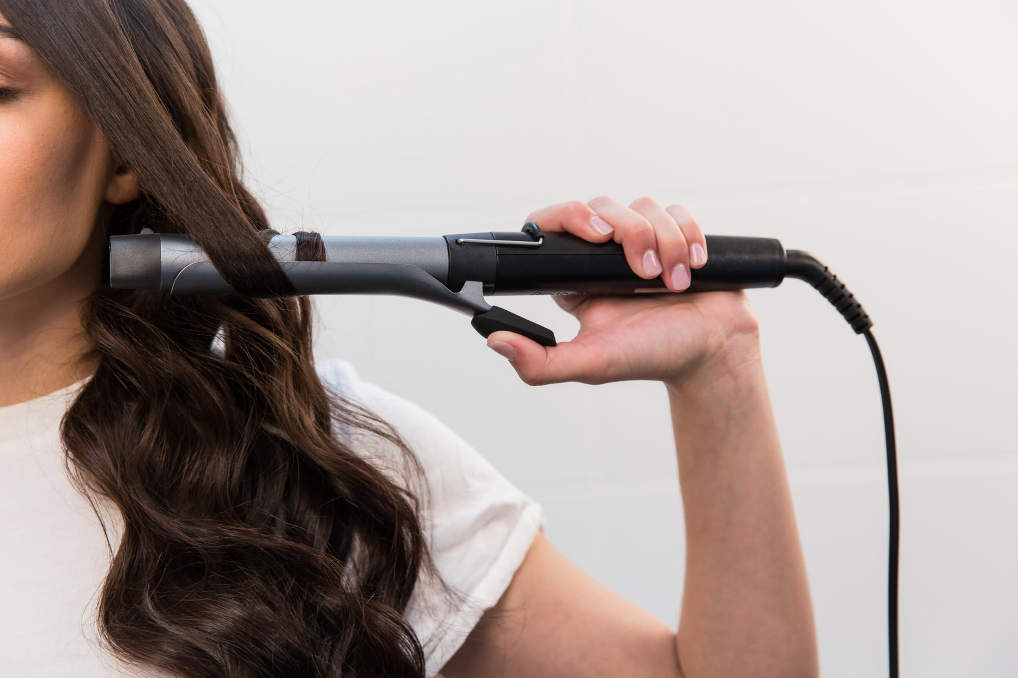 Donna che si arriccia i capelli con un ferro arricciacapelli. Il ferro è grigio e i capelli sono castano scuro.