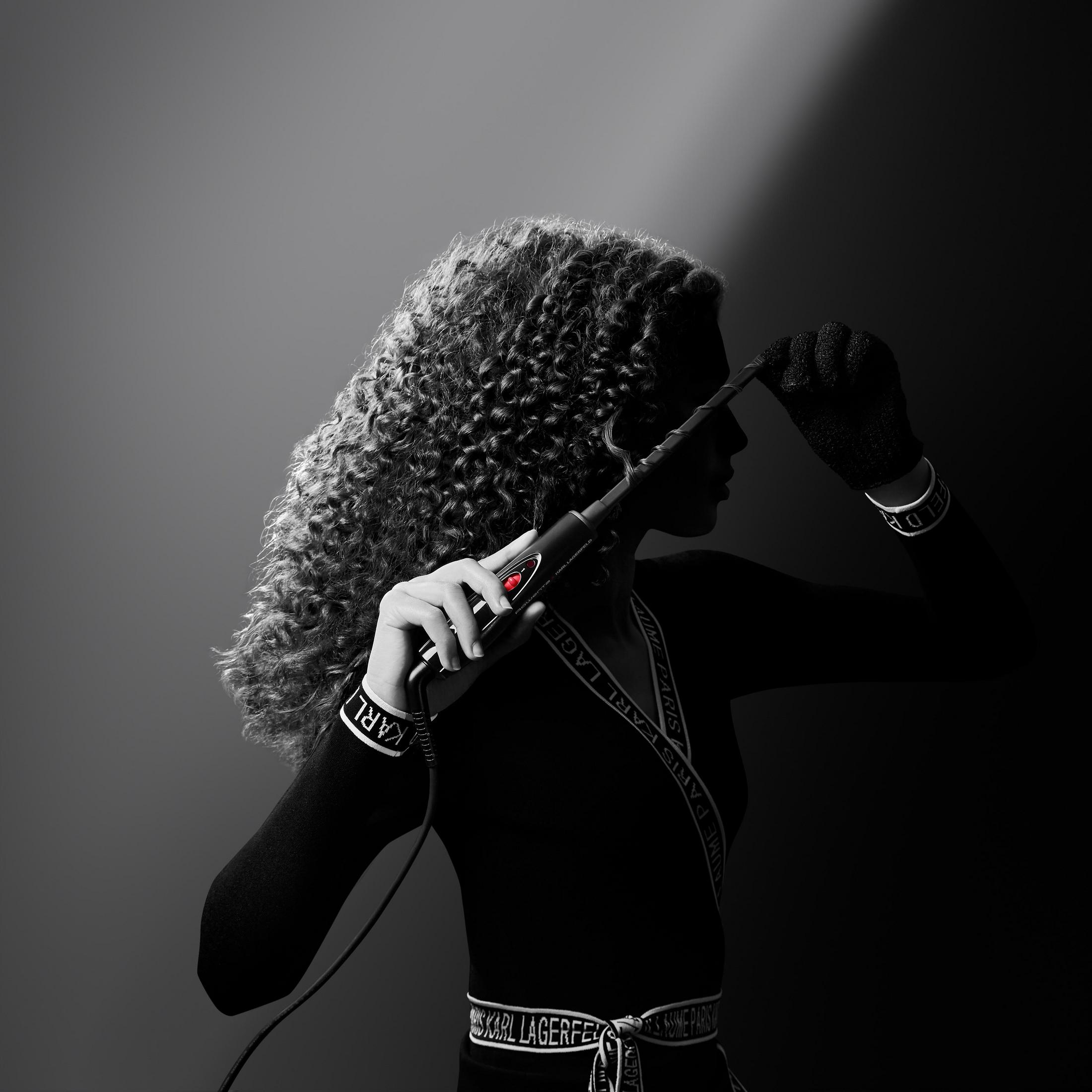 Kobieta z kręconymi włosami używająca lokówki, w czarnych ubraniach, na szarym tle.