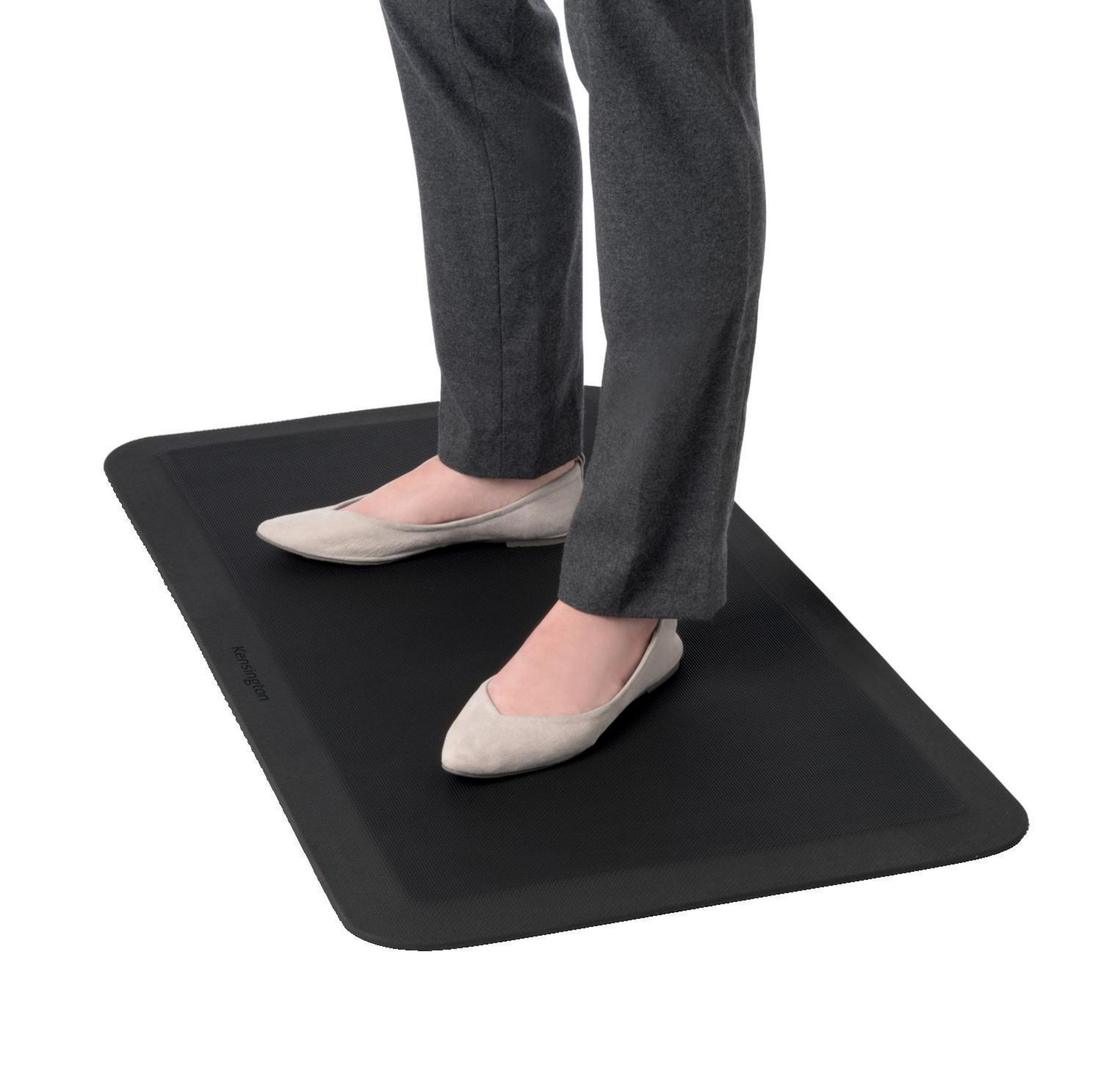 Person steht auf einer schwarzen, rechteckigen Anti-Ermüdungsmatte. Graue Hose, beige Schuhe.