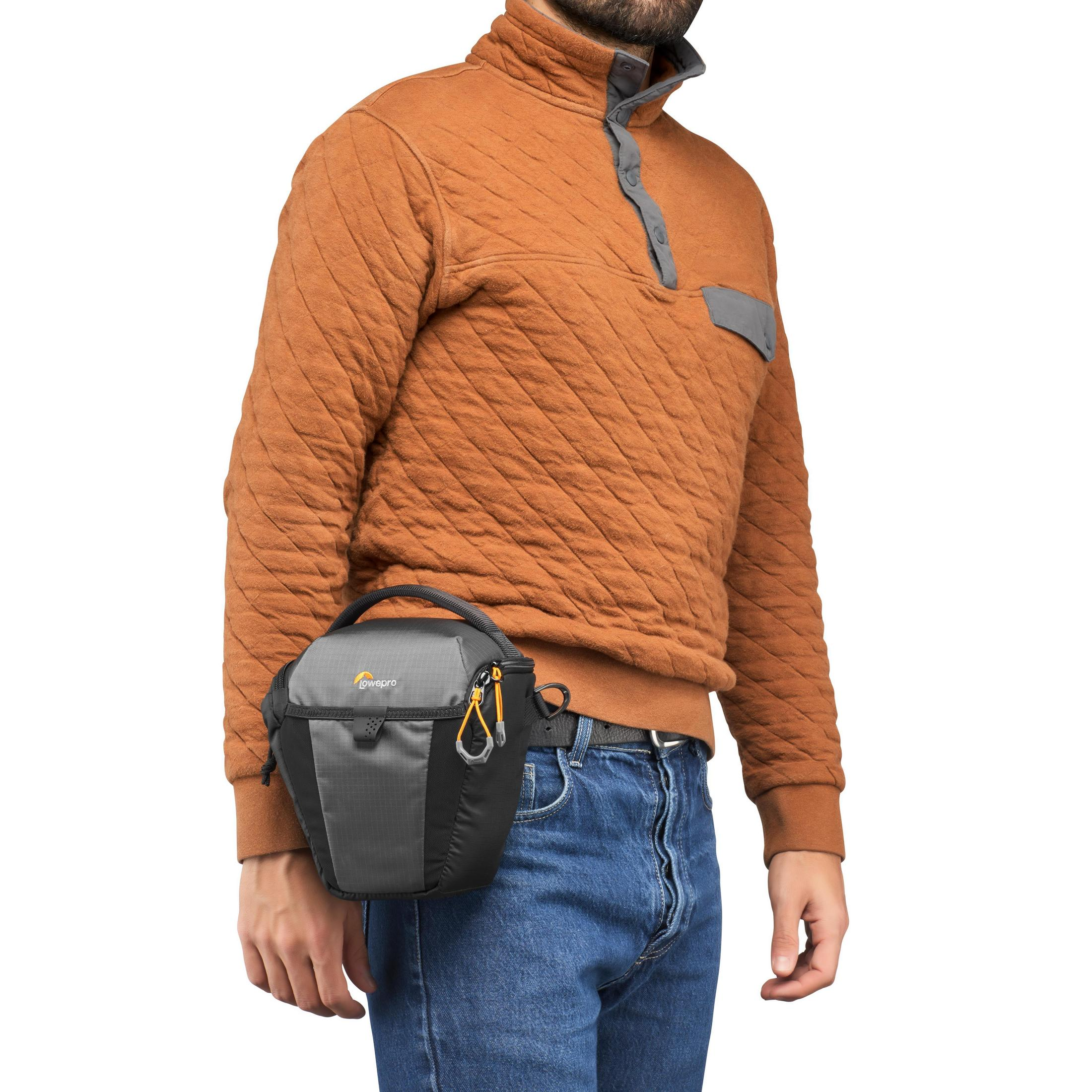 Ein Mann mit braunem Pullover und Kameratasche um die Taille, hält eine Kamera.