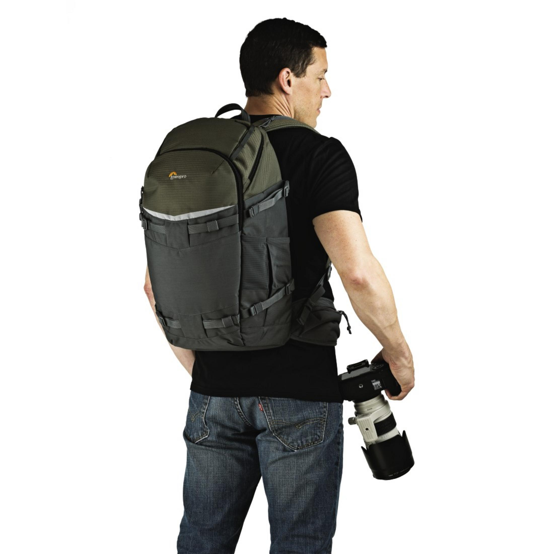 Un homme portant un sac à dos, tenant un appareil photo et un objectif, sur fond blanc.