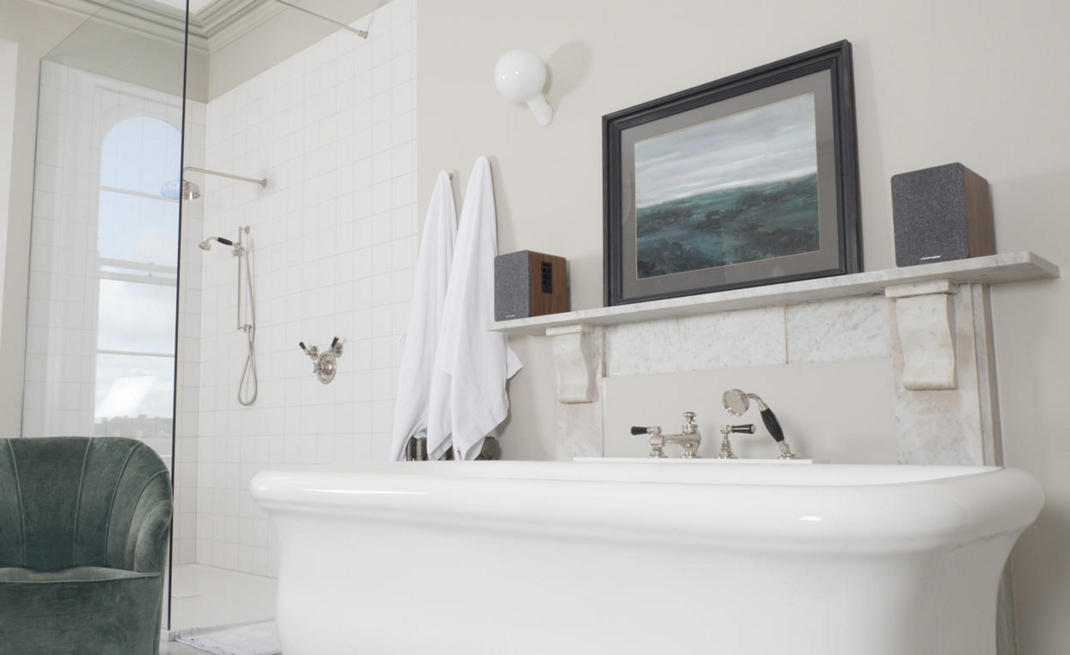 Badezimmer mit weißen Fliesen, Badewanne, Dusche und Kunst. Lautsprecher und Handtücher vorhanden.