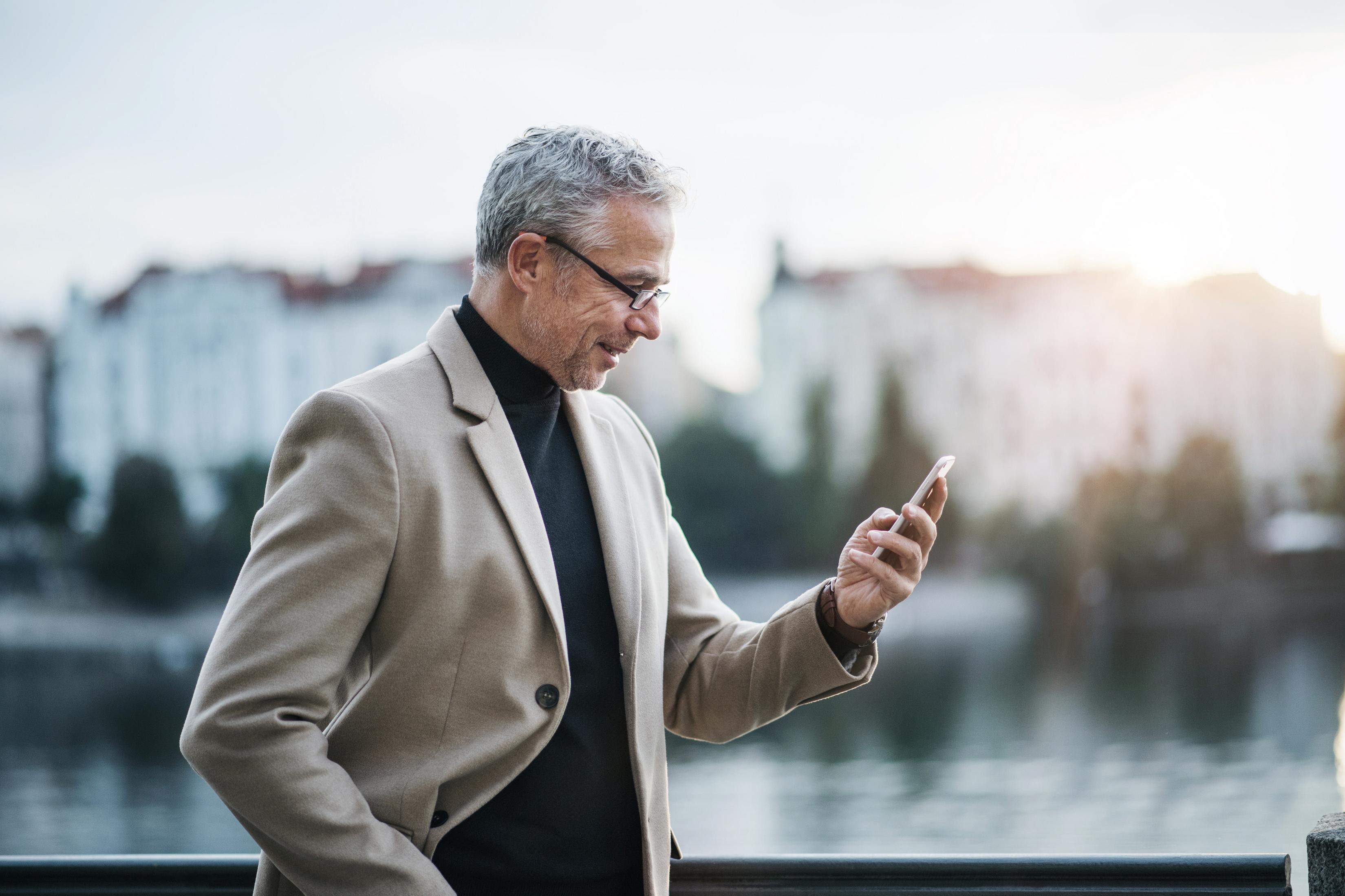Mann mit Brille schaut auf sein Handy. Er trägt eine Jacke und steht draußen.
