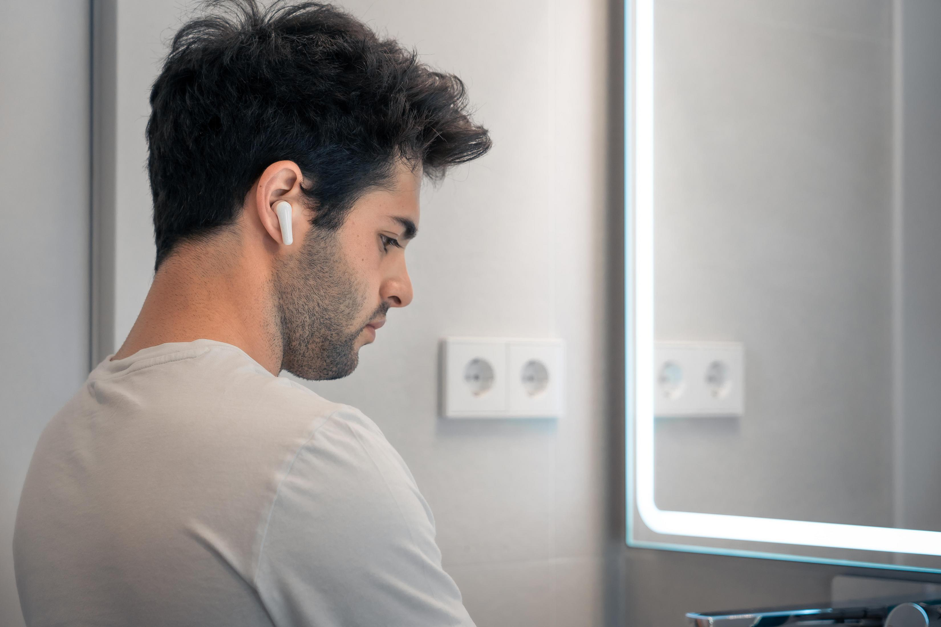 Man in a white shirt wearing a white earbud, looking at a mirror.