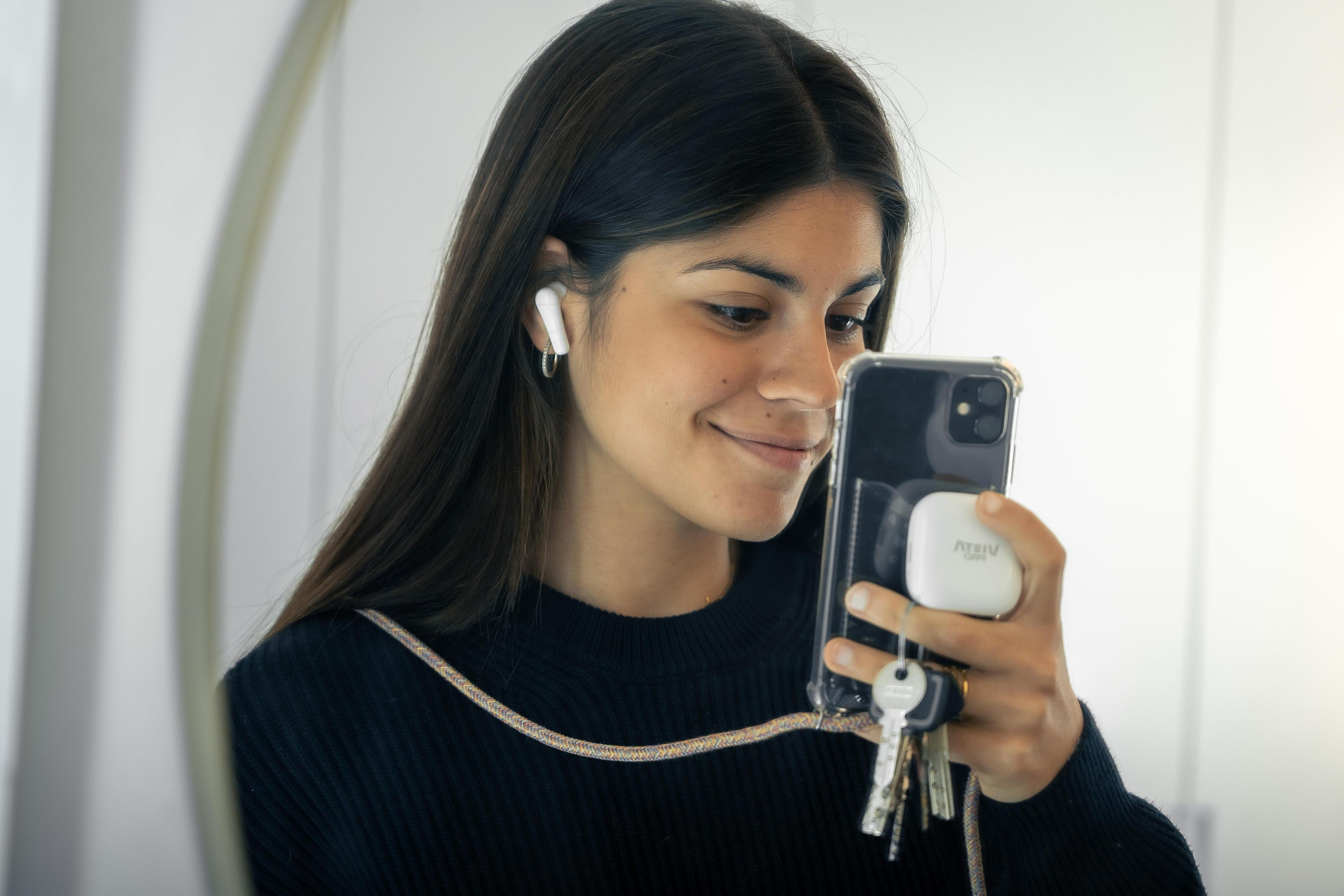 A woman smiles while looking at her phone. She wears earbuds and holds a phone with keys attached.