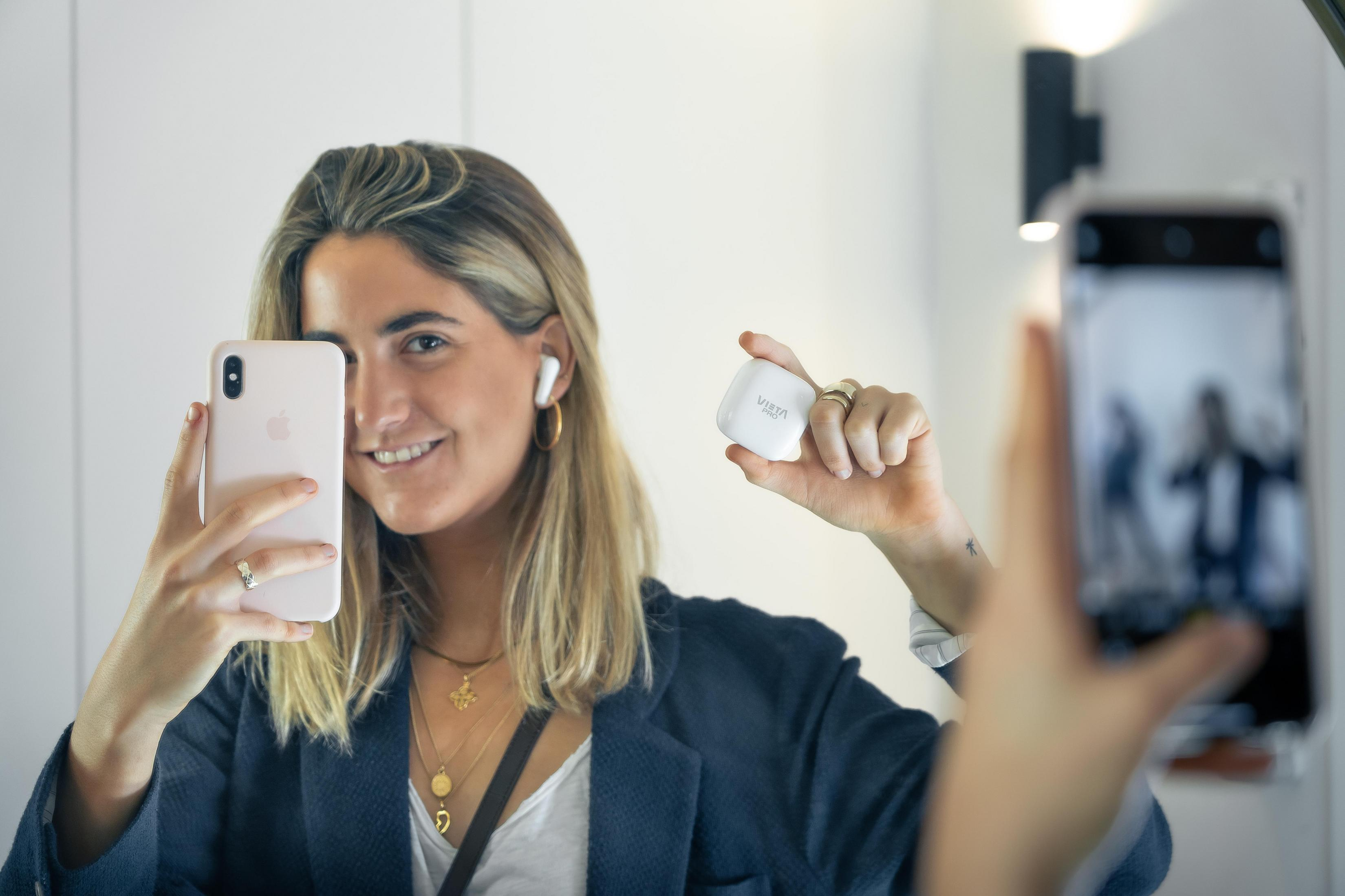 A woman is taking a selfie with a pink phone and holding a white earbud case, smiling at the camera.