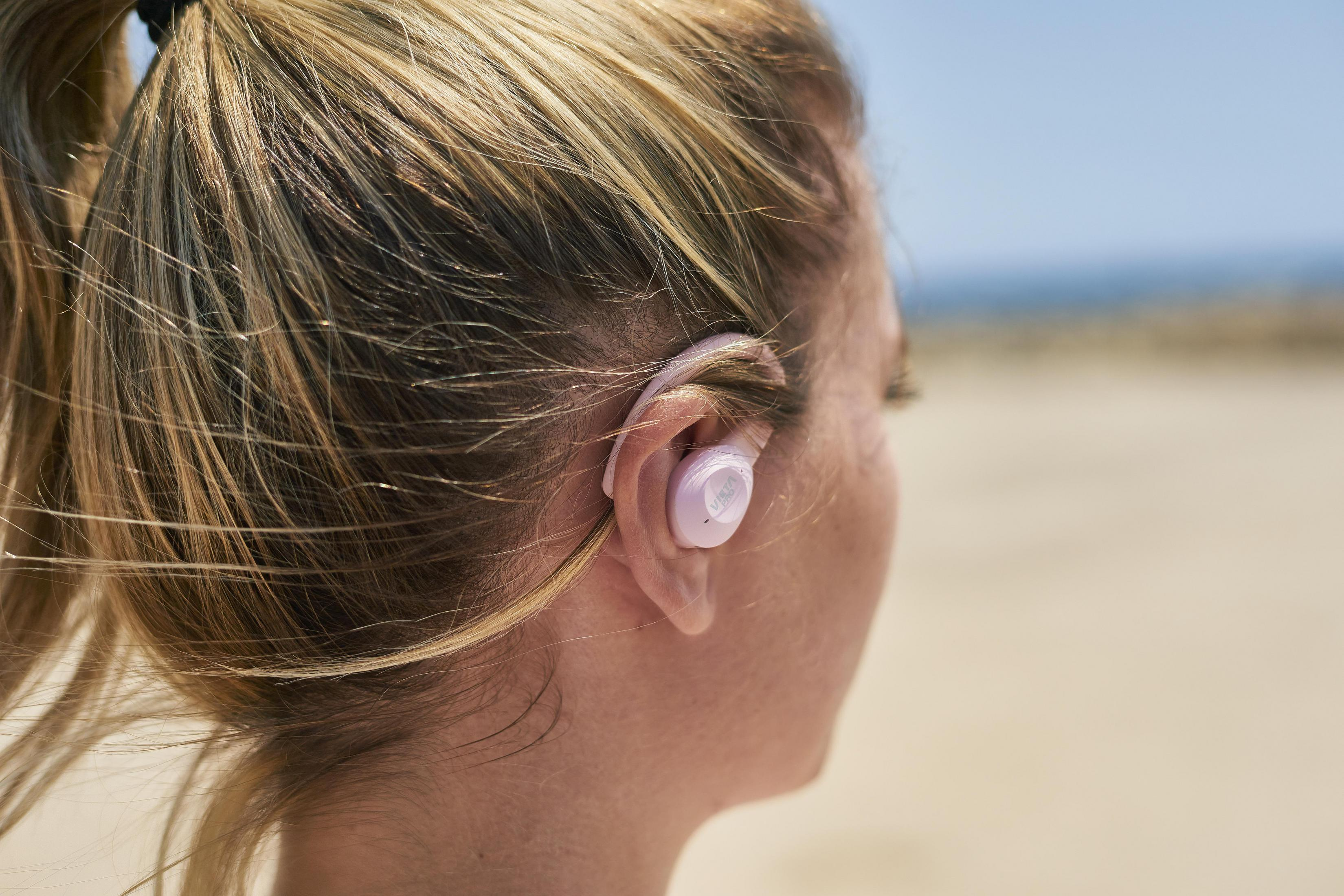 Donna con un auricolare wireless rosa. Lo sfondo è una scena sulla spiaggia.