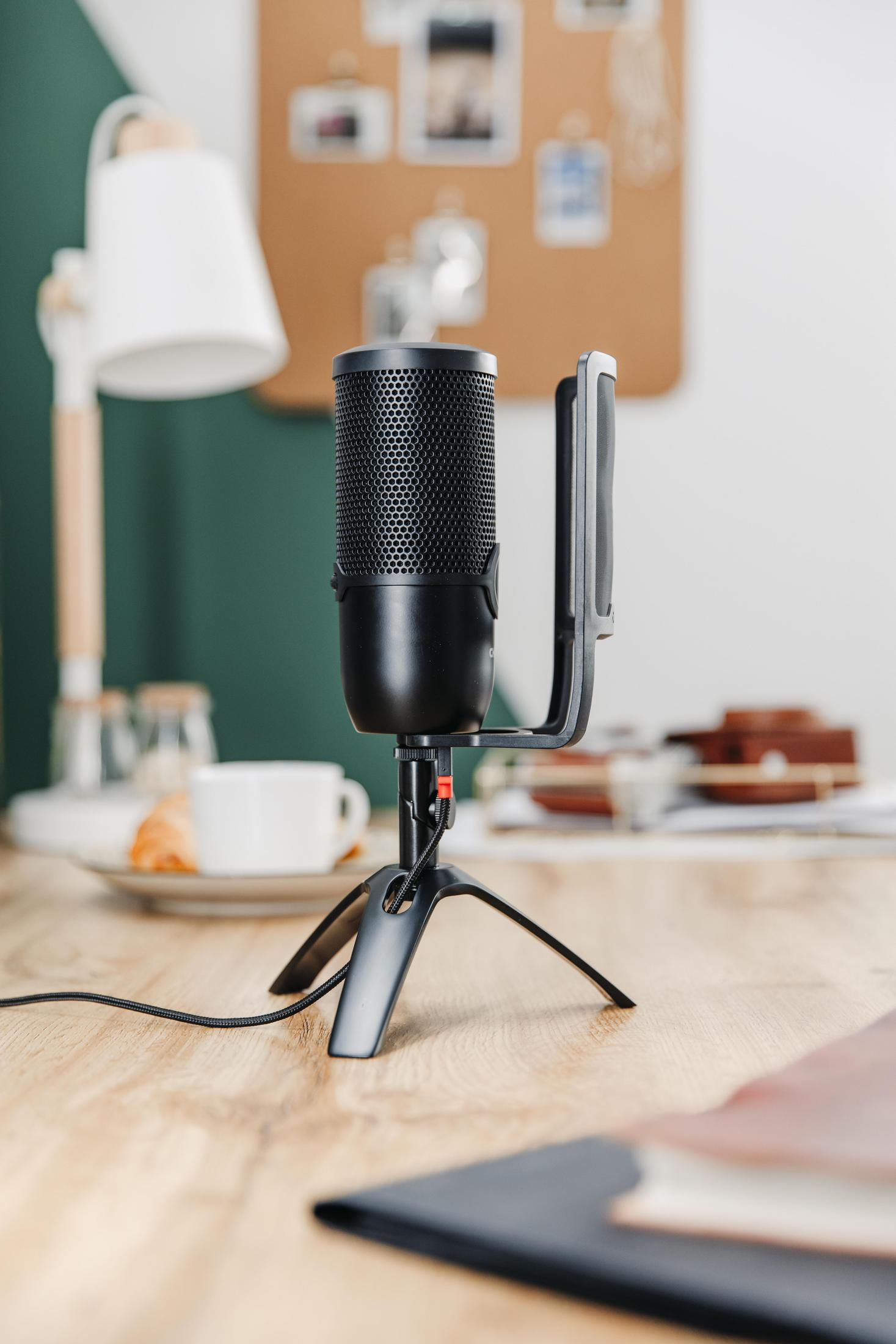 Schwarzes Mikrofon auf Ständer. Schreibtisch mit Kaffee, Lampe und Pinnwand.
