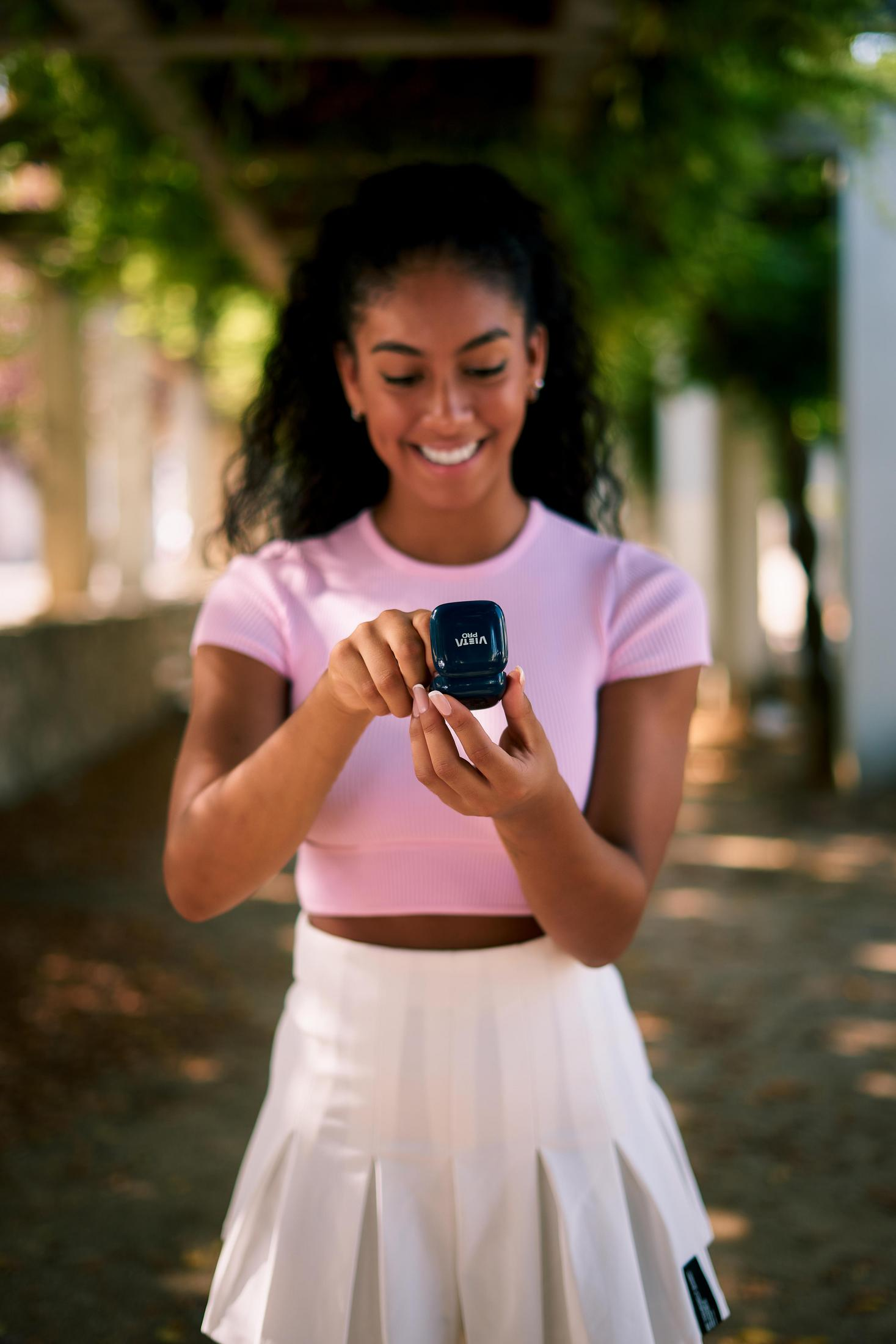 A woman in pink top and white skirt smiles, holding a small black device outdoors.