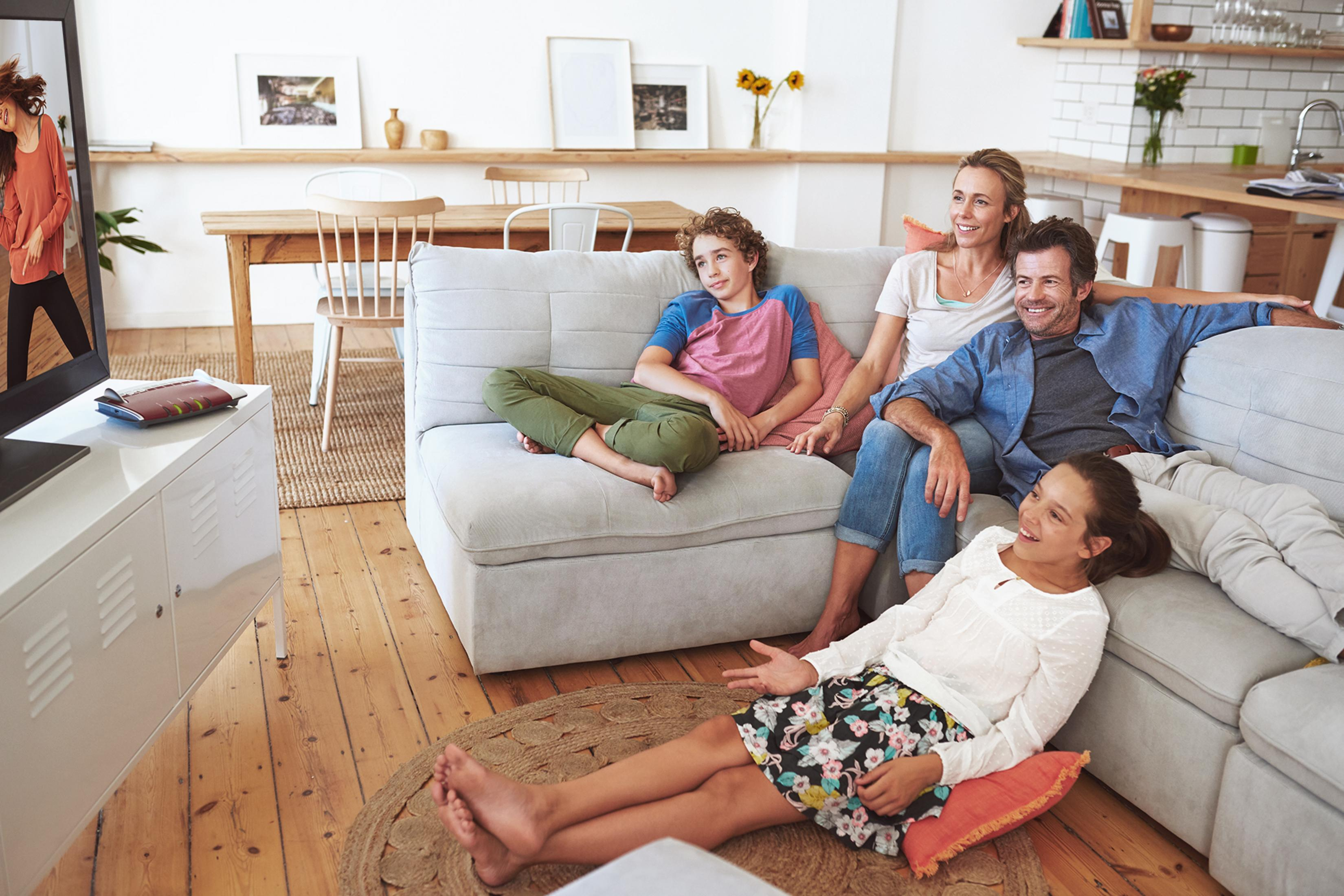 Familie schaut fern auf dem Sofa. Der Raum hat Holzböden und eine weiße Wand.