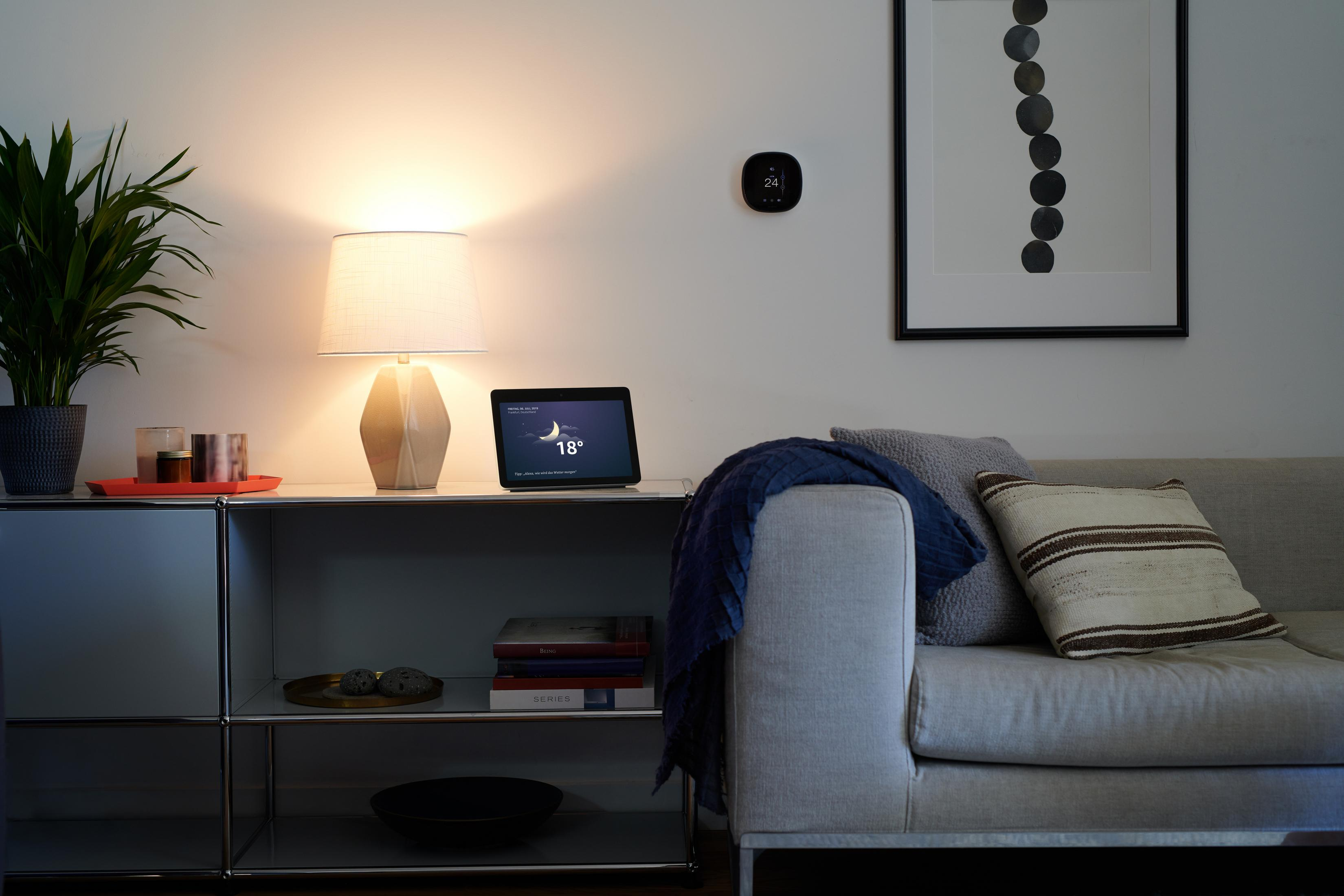 Ein Raum mit Lampe, Tisch und intelligentem Display mit Wetteranzeige. Sofa und Kunst.