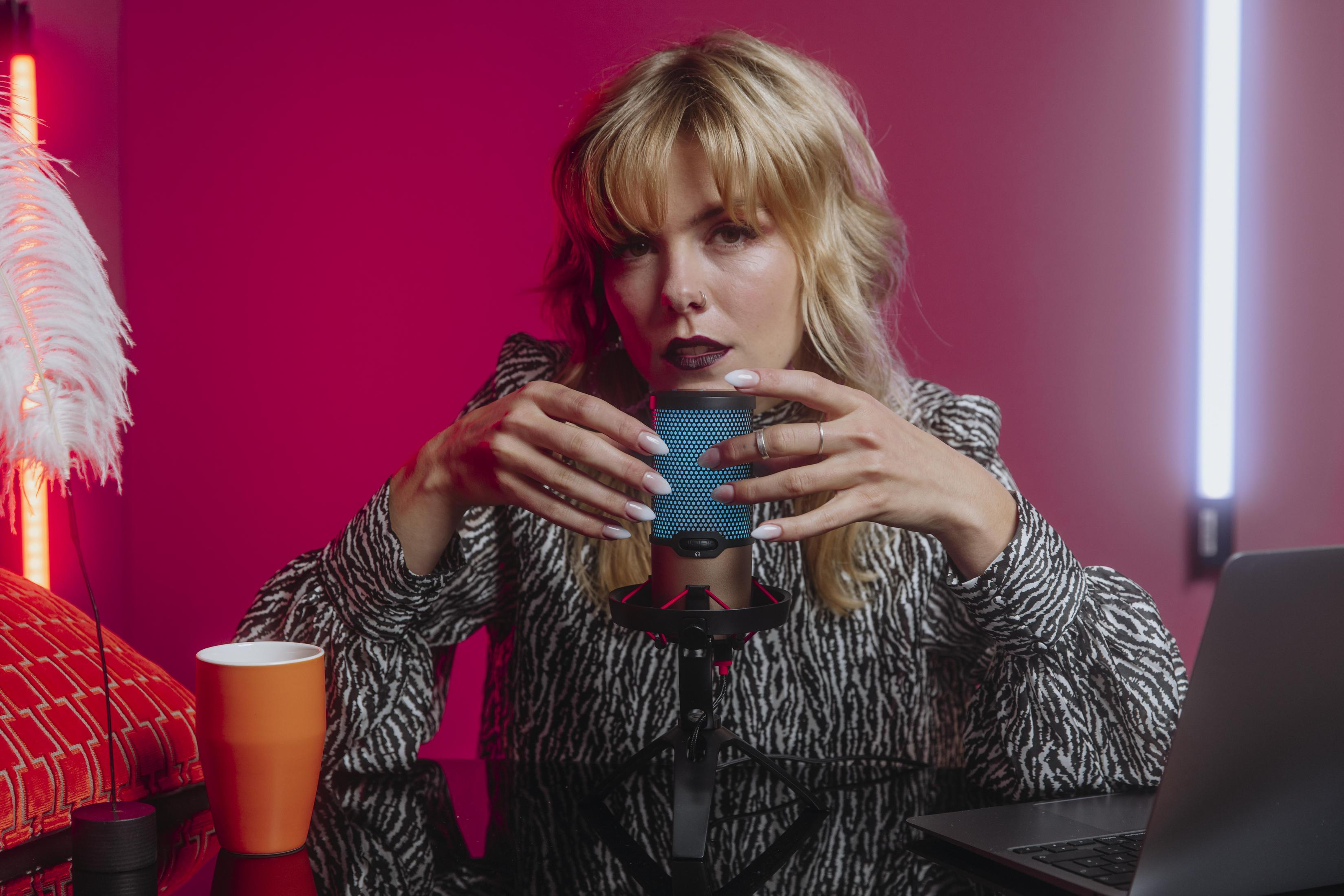 Woman recording with a microphone. There's a coffee mug, laptop, and pink background.