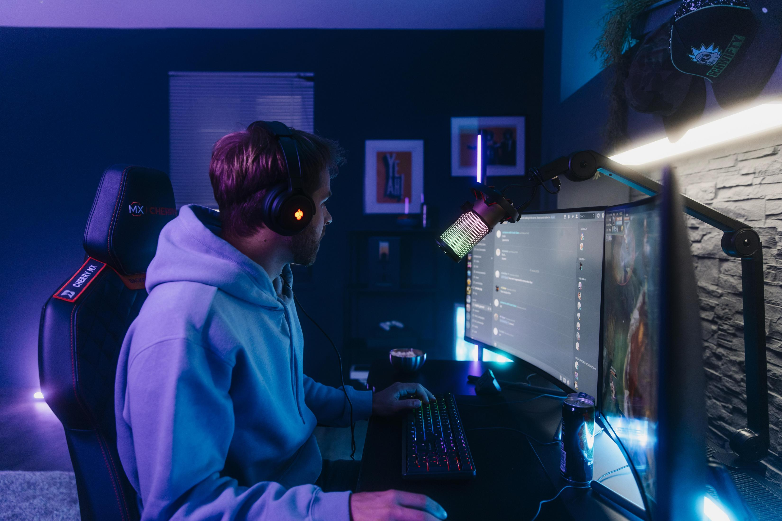 A man wearing headphones gaming on a computer with a microphone in a dark room.