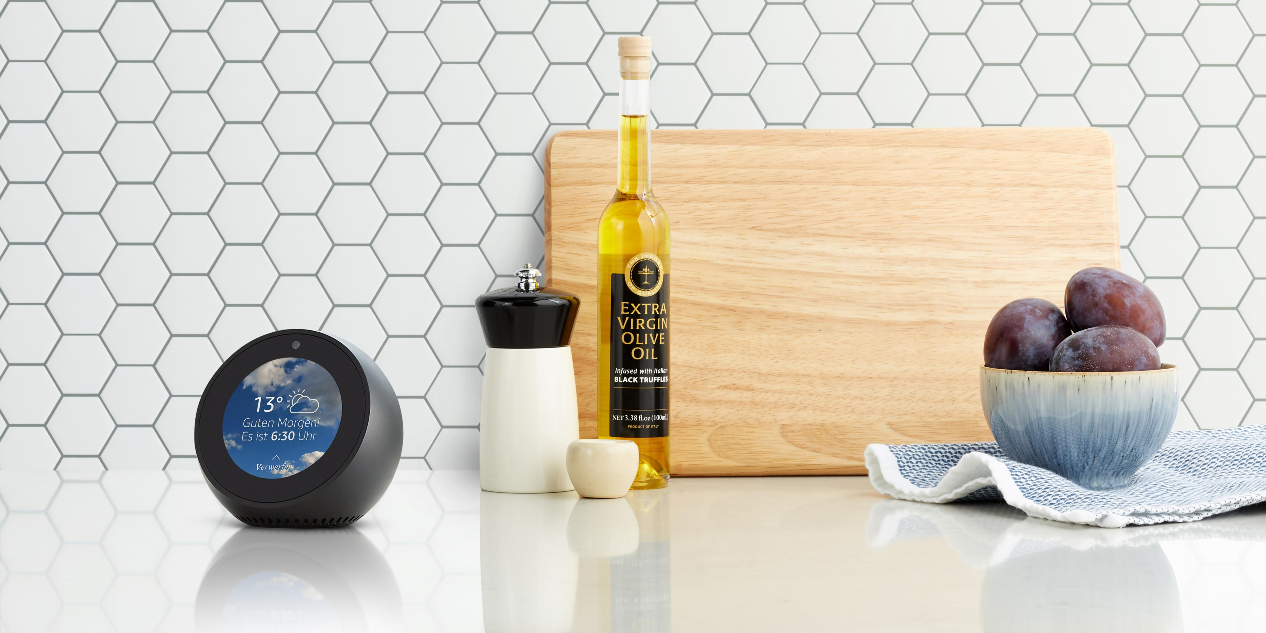 Ein Echo Show zeigt die Wettervorhersage. Olivenöl, Pflaumen und ein Schneidebrett.