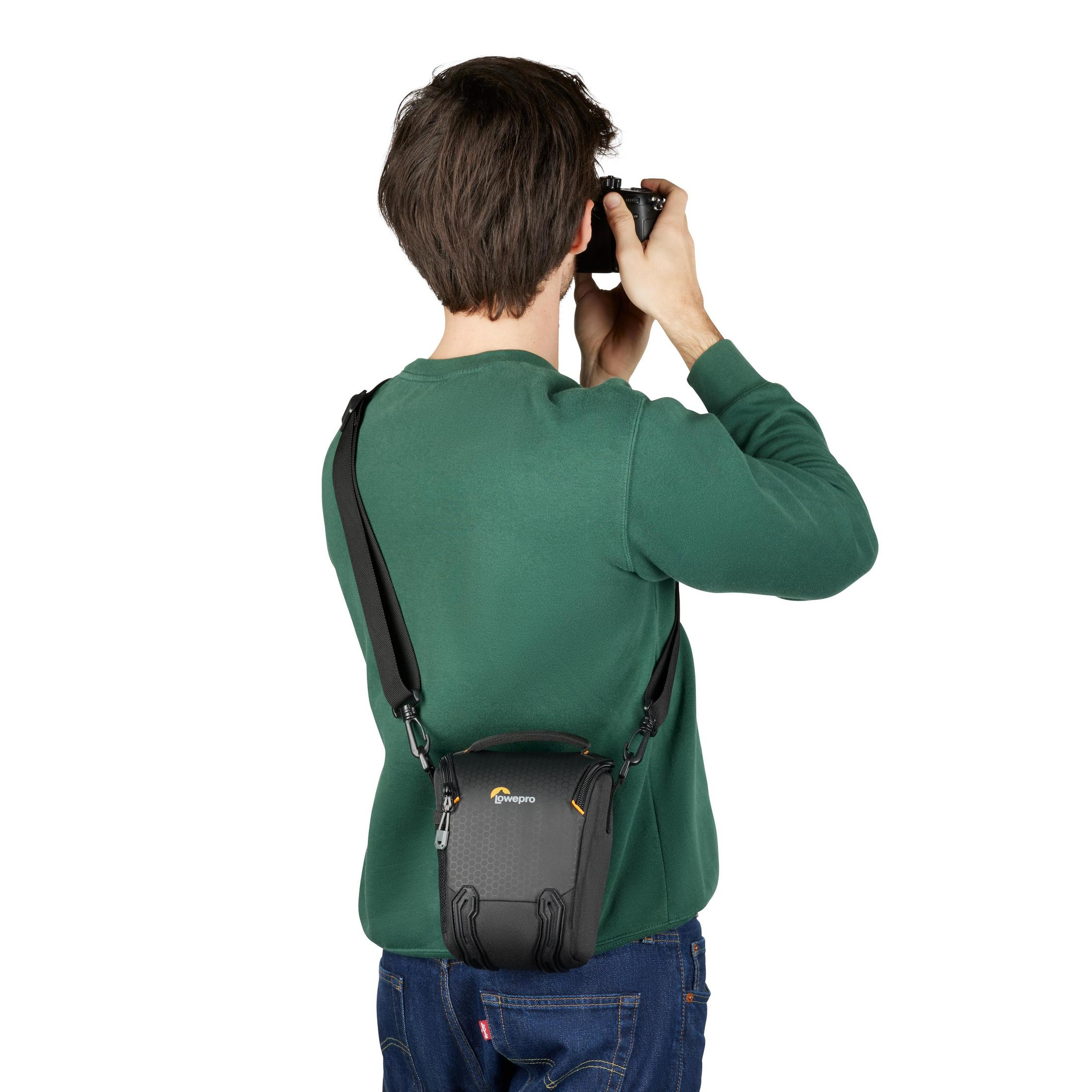 Person taking a photo with a camera. The person wears a green shirt, camera bag is visible.