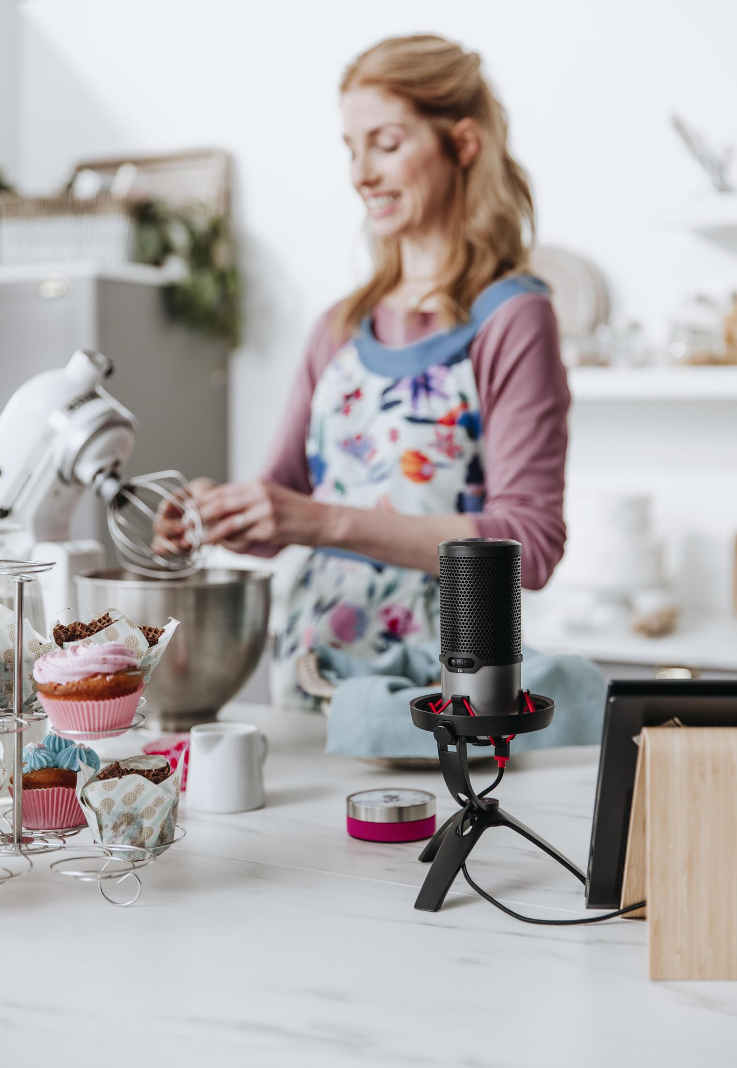 Vrouw in keuken, beslag maken. Cupcakes vooraan. Microfoon op standaard staat ook vooraan.