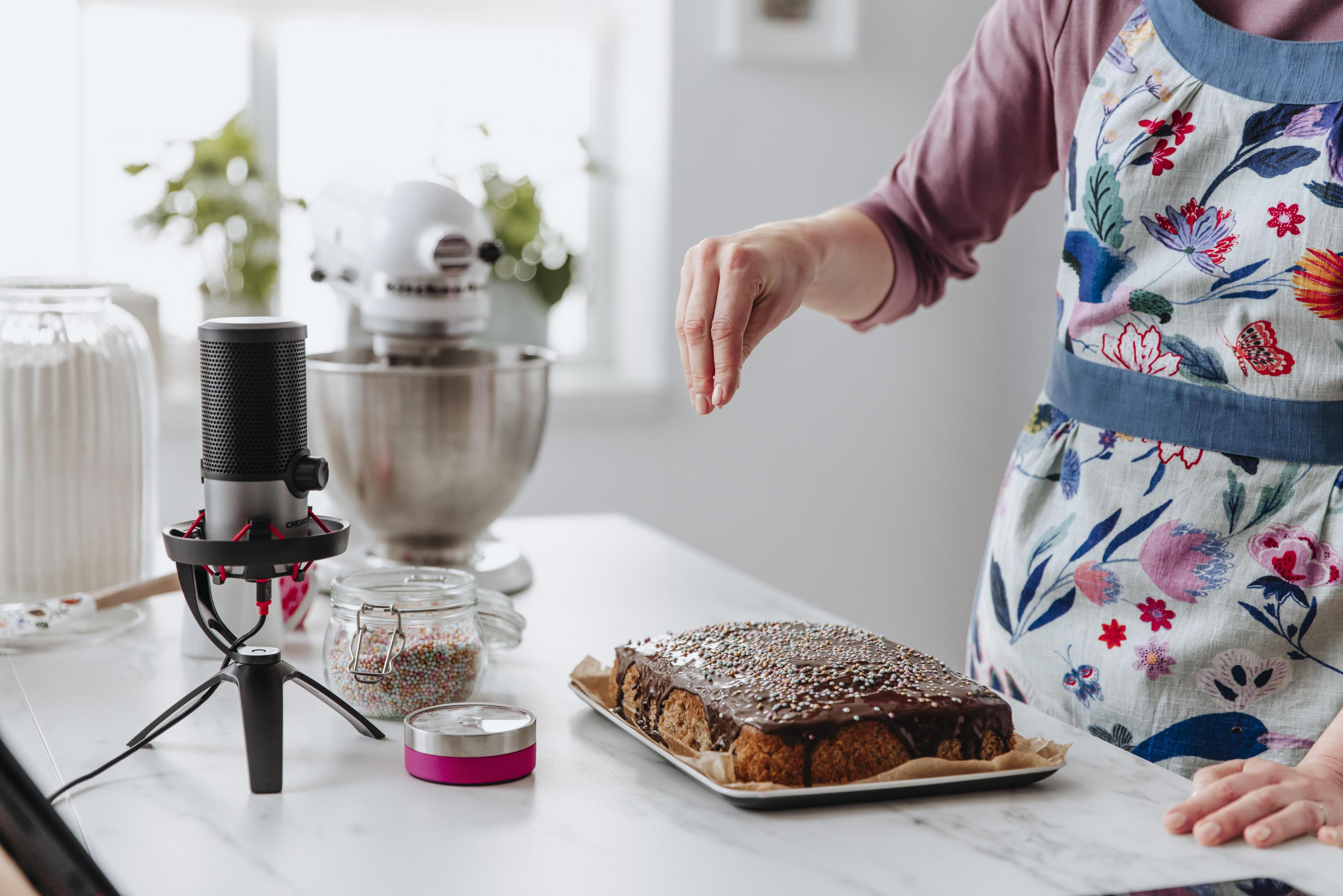 Een vrouw strooit hagelslag op een met chocolade bedekte cake. Een microfoon en een mixer staan op tafel.