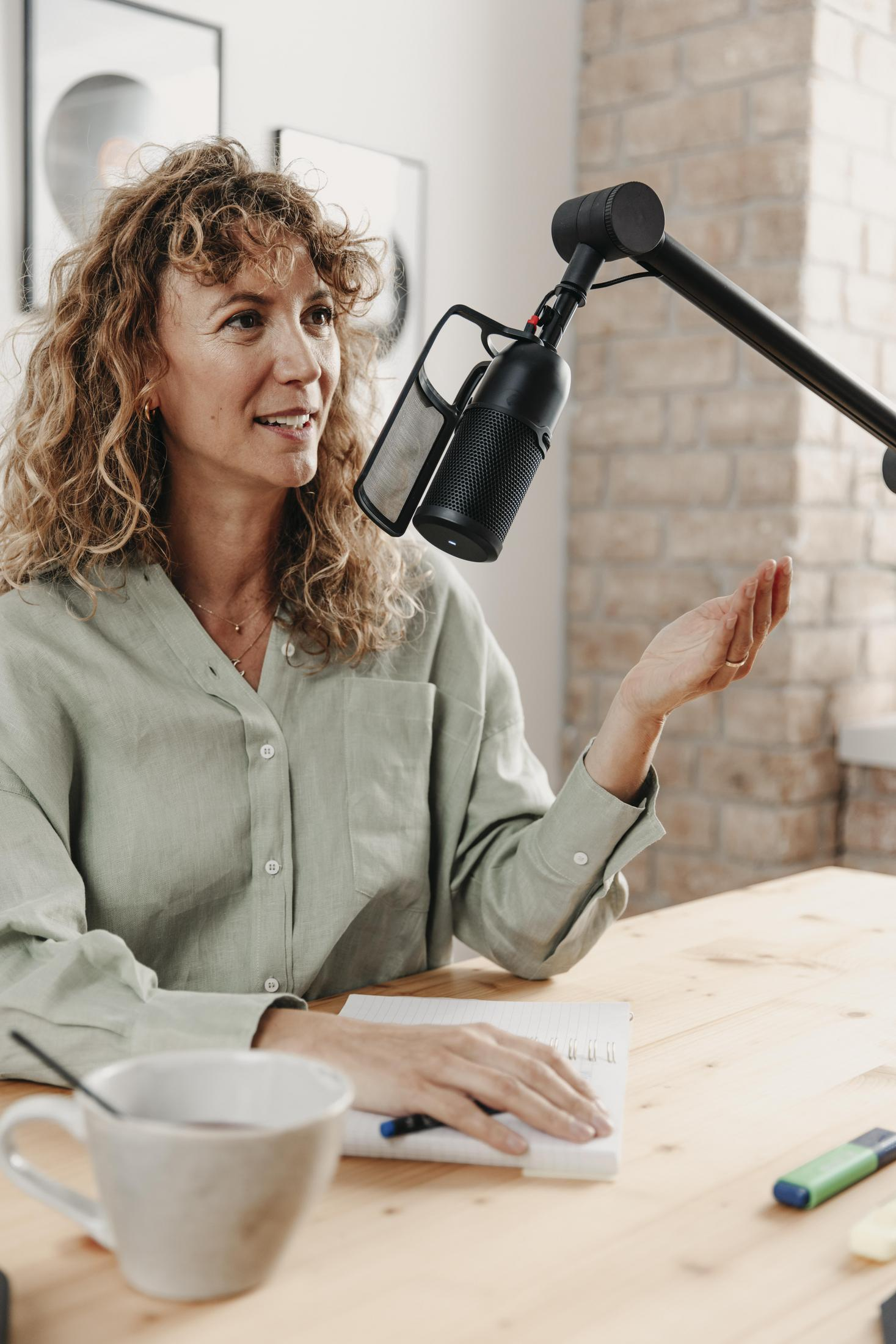 Een vrouw spreekt in een microfoon met een popfilter. Ze zit aan een bureau met een notitieboek en een kopje.