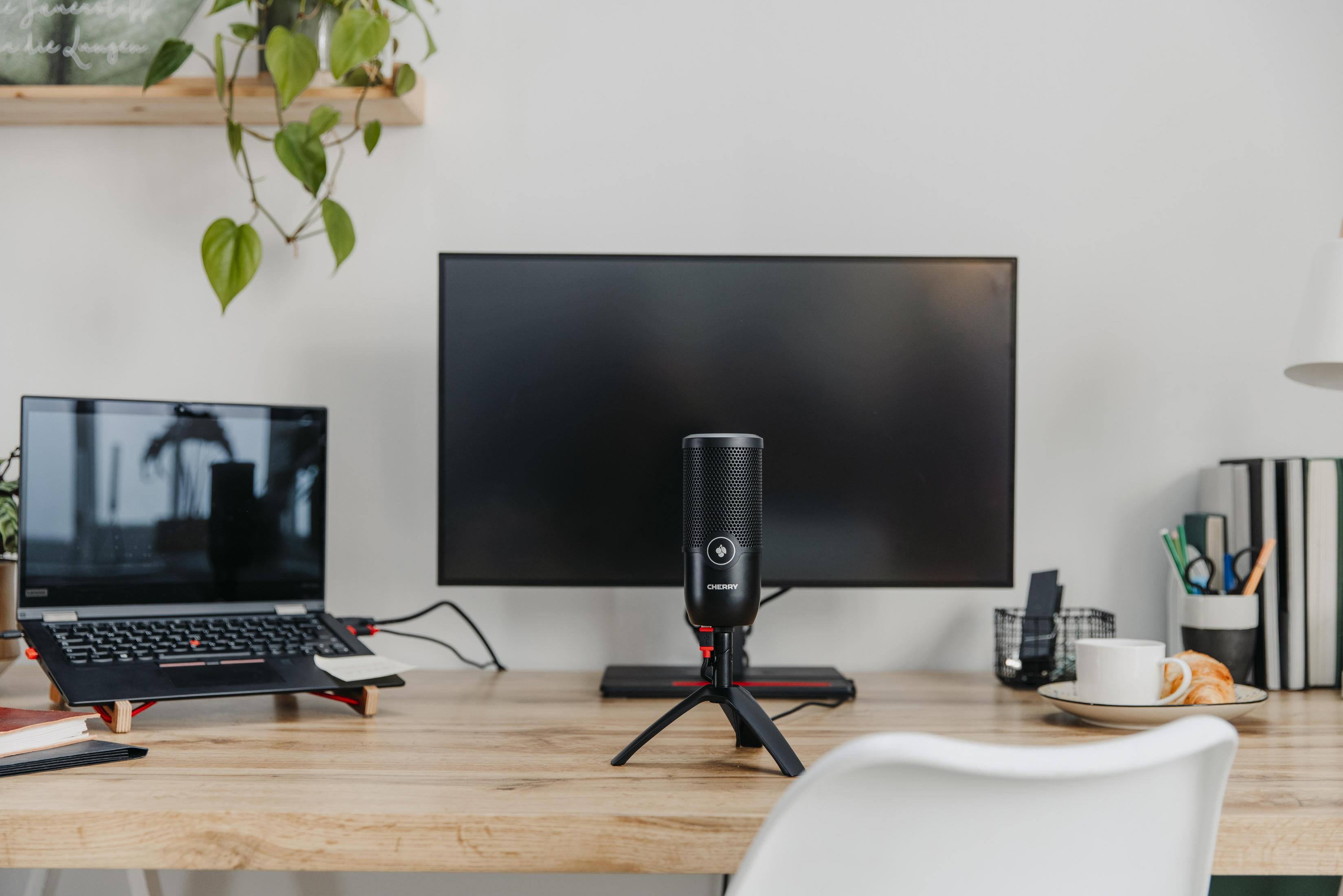 Een bureau met een laptop, microfoon, monitor en andere kantoorbenodigdheden. Een kopje koffie en een croissant liggen op het bureau.