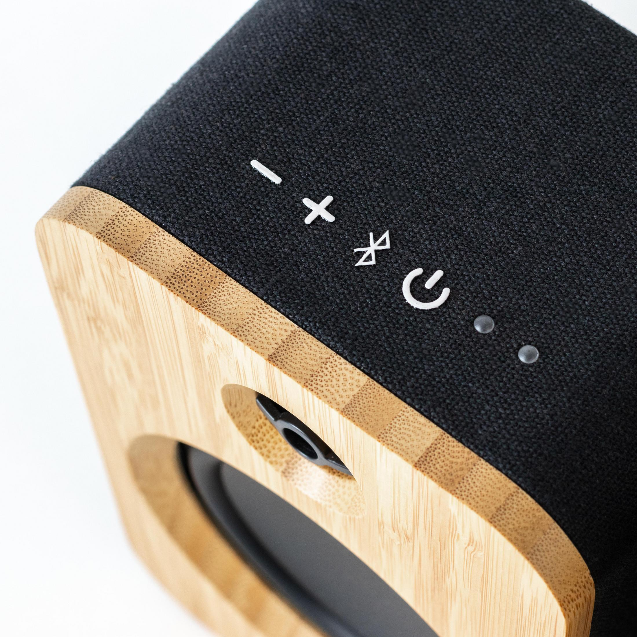 Haut-parleur en bois avec dessus en tissu noir. Les commandes incluent moins, plus, bluetooth et alimentation.