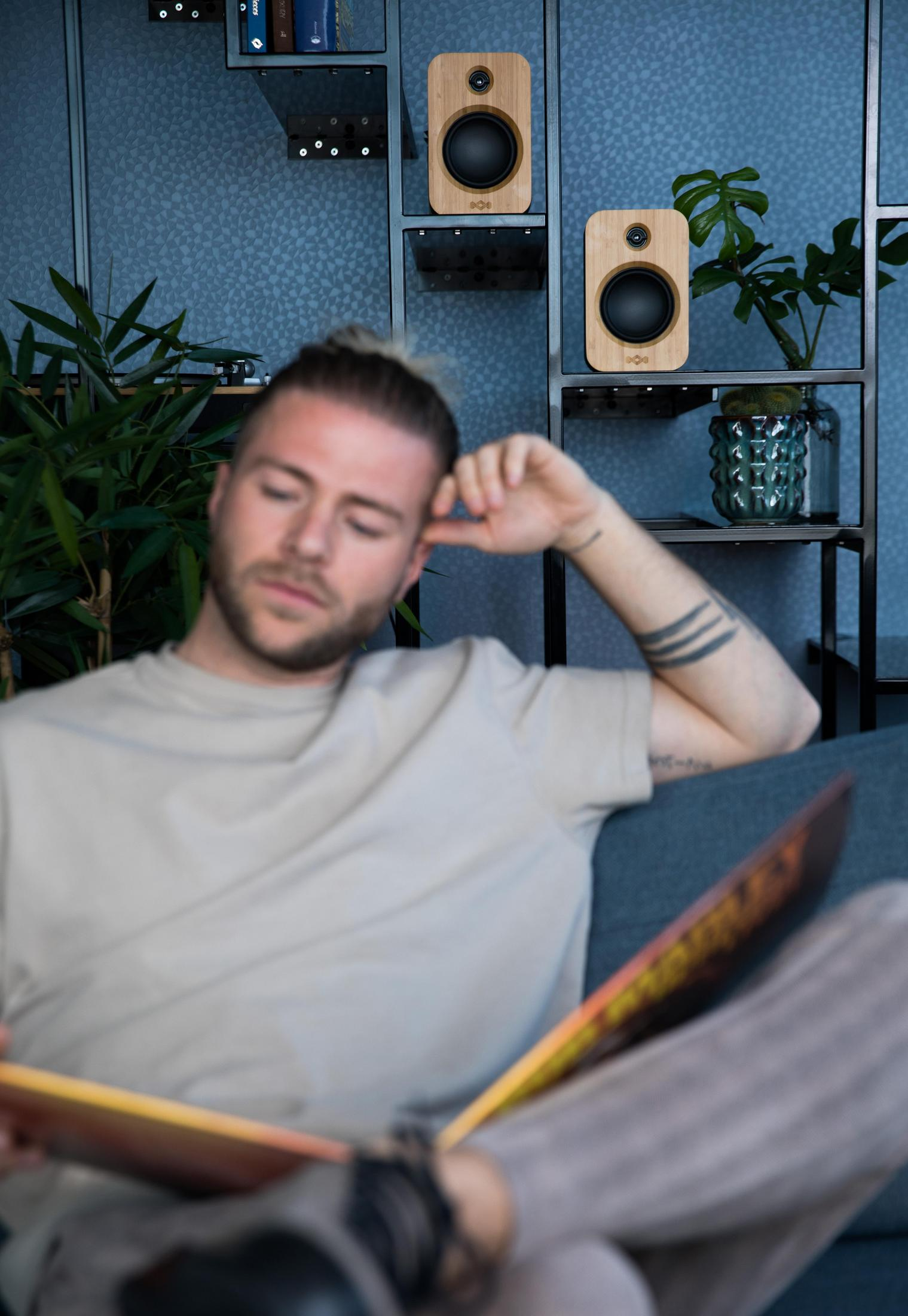 Homme lisant un livre, assis sur un canapé, avec des enceintes et une plante en arrière-plan.
