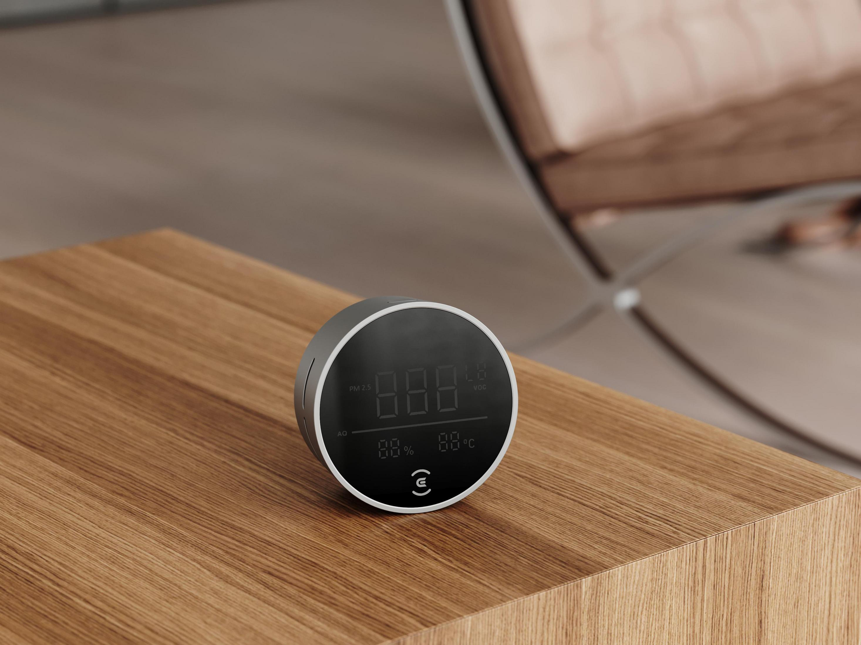 Luftqualitätsmonitor auf einem Holztisch. Das Display zeigt Zahlen, mit einem Sessel im Hintergrund.