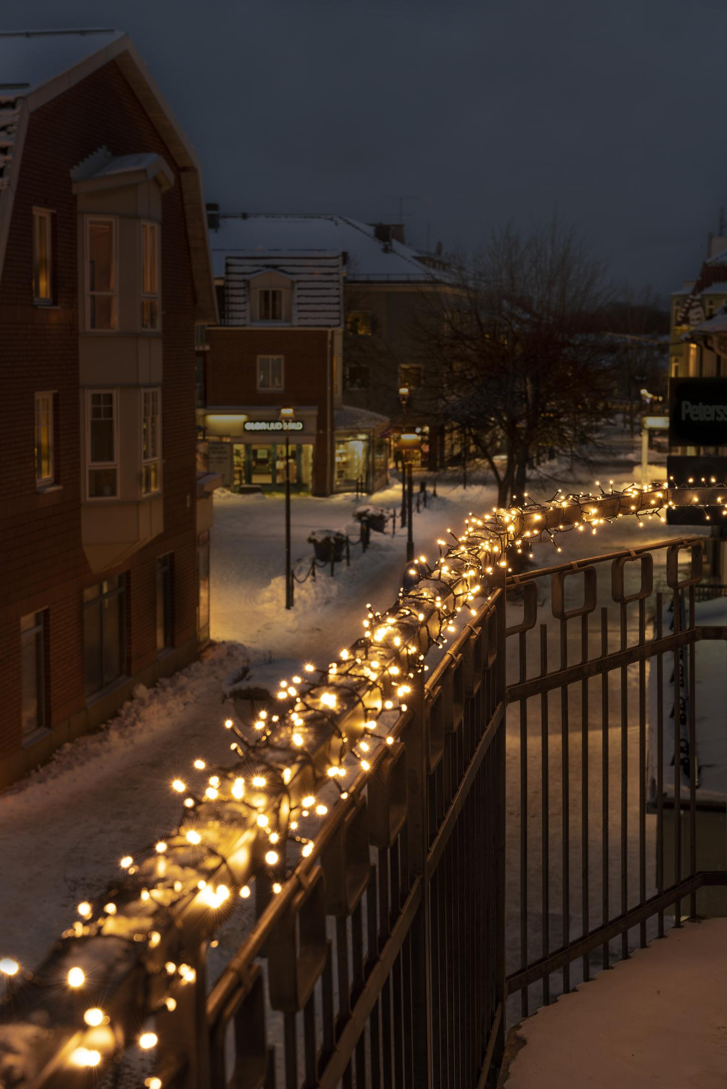 Nachtszenen mit verschneiter Straße, Gebäuden und einem Geländer mit Lichterketten.