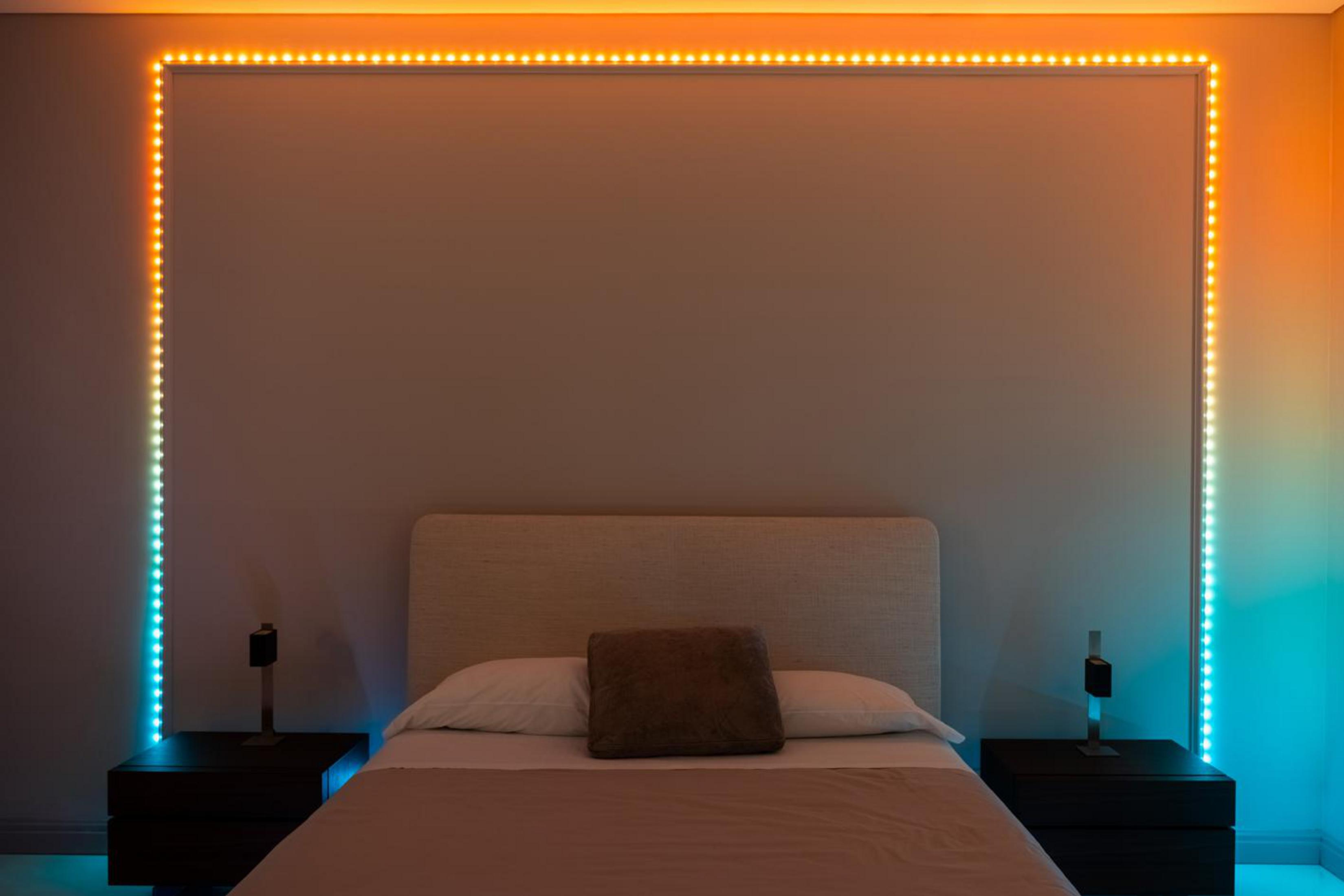 Łóżko z szarą ramą, dwoma szafkami nocnymi i kolorowymi światłami LED na ścianie.