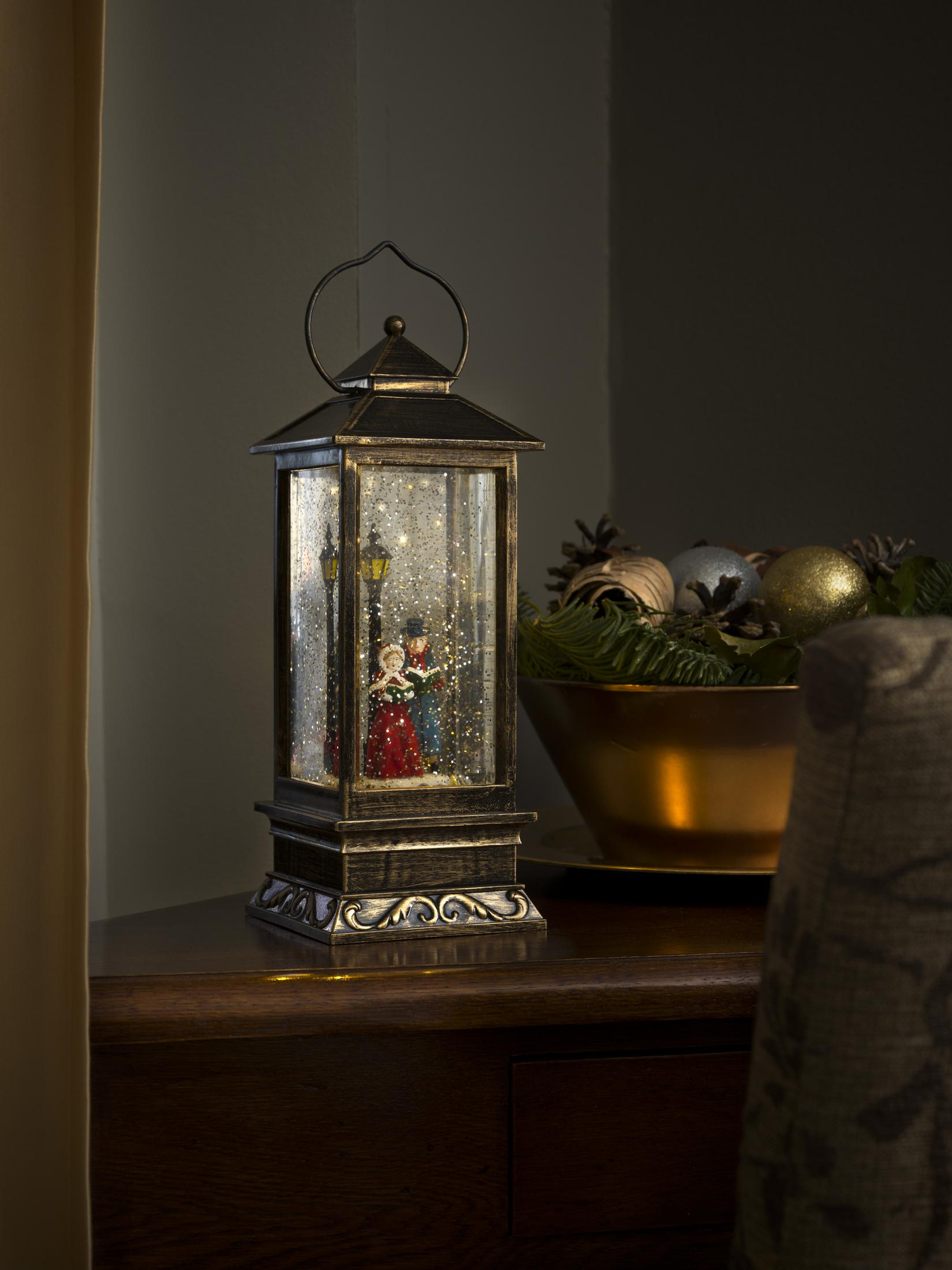 Eine Weihnachtsszene in einer Laterne mit Schnee. Paar und Laternenpfahl darin. Schale mit Ornamenten im Hintergrund.