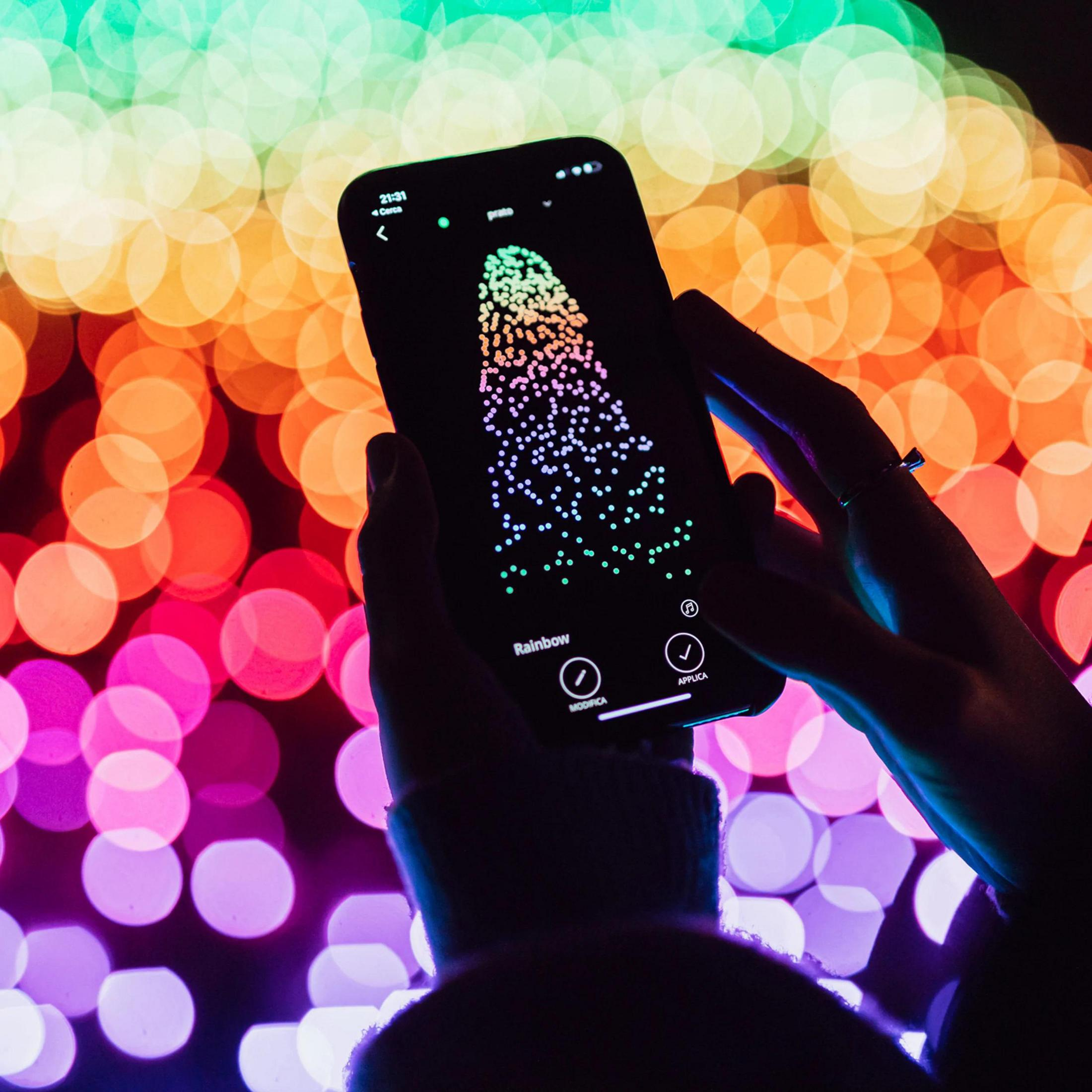 Ekran smartfona pokazuje aplikację w kolorach tęczy, trzymaną w dłoniach na tle rozmytych świateł.