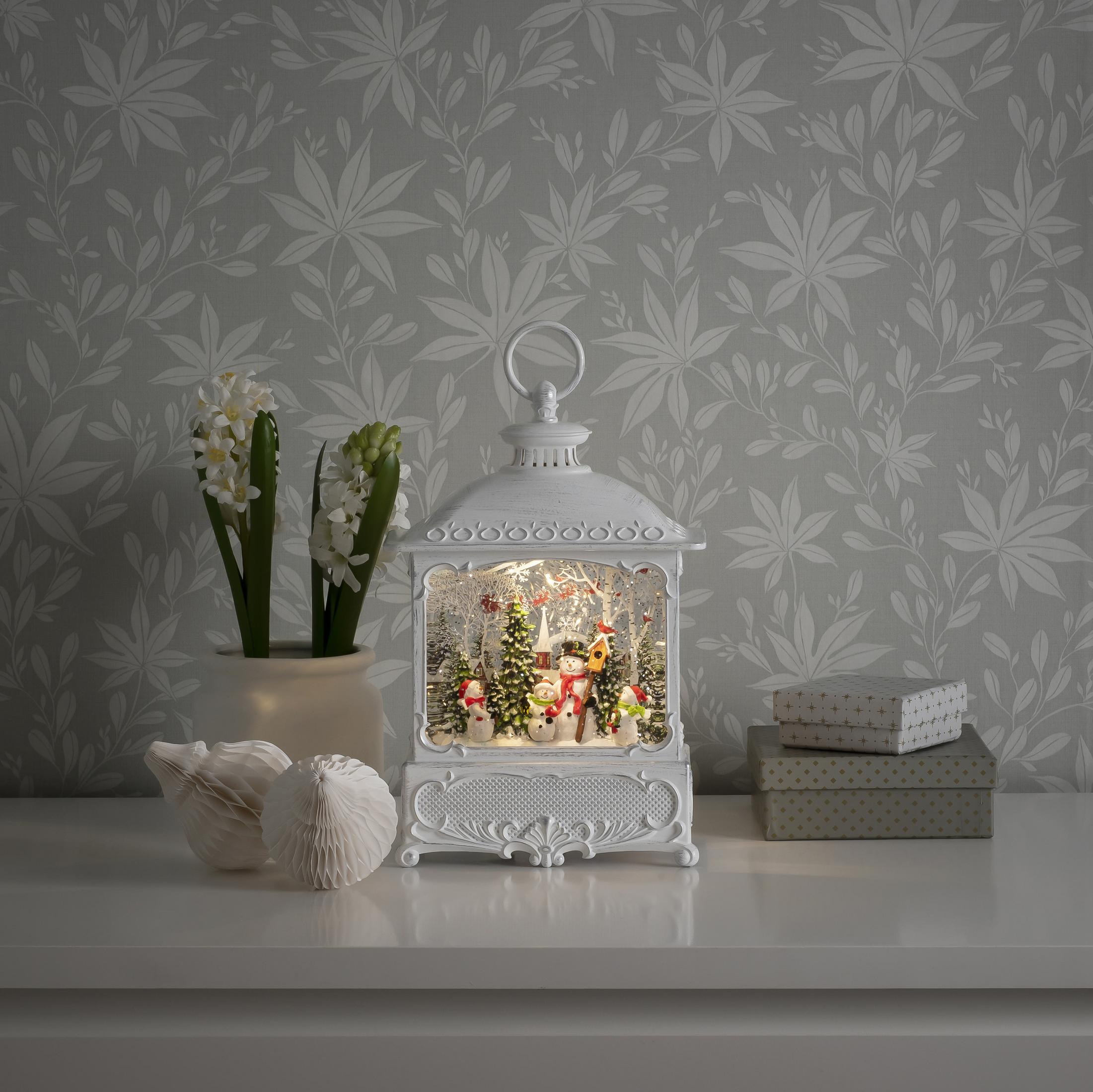 Weiße Laterne mit Weihnachtsszene, auf weißem Tisch. Weiße Blumen und Geschenke.