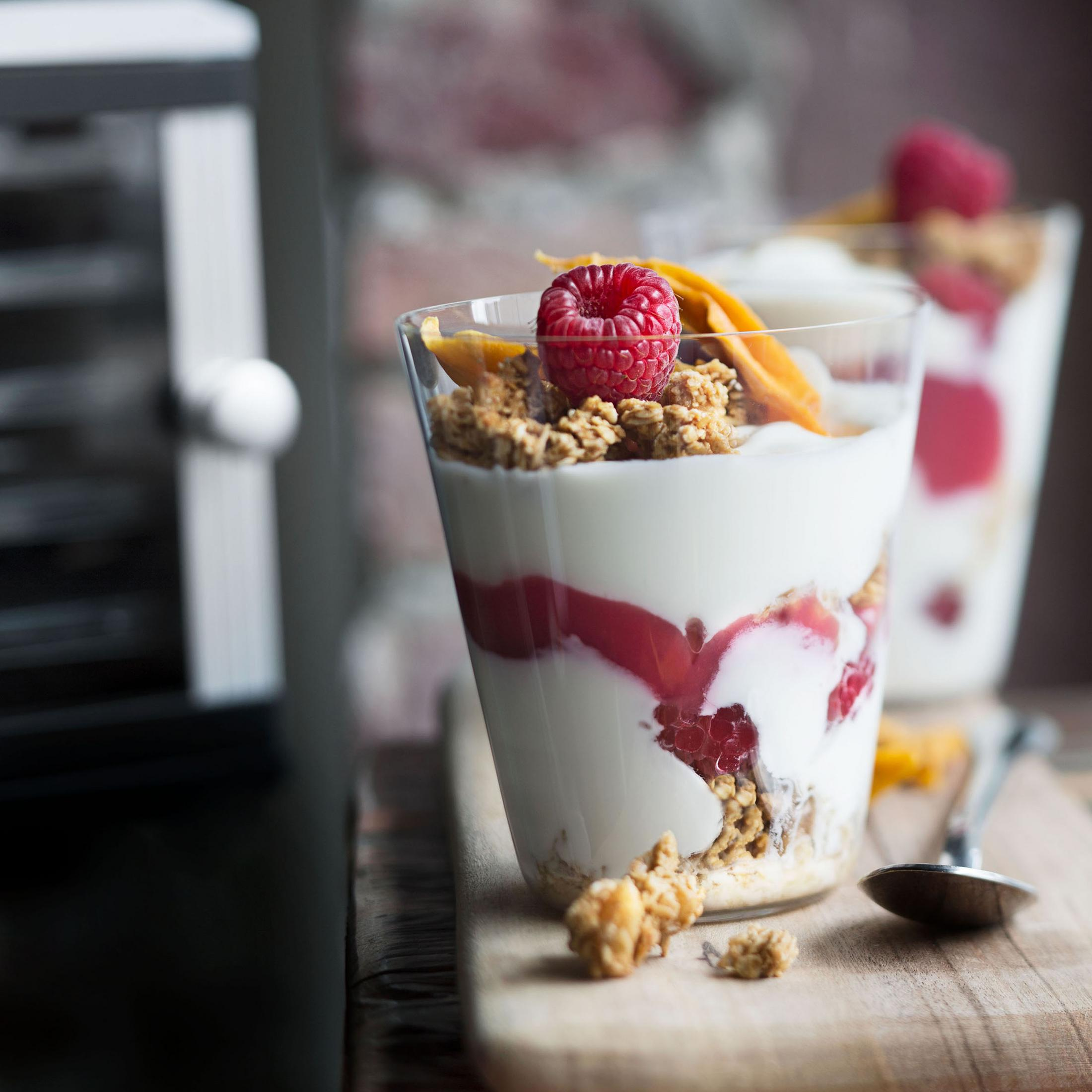 Zwei Gläser Joghurt-Parfait mit Granola, Himbeeren und einer Mangoscheibe auf einem Holzbrett.