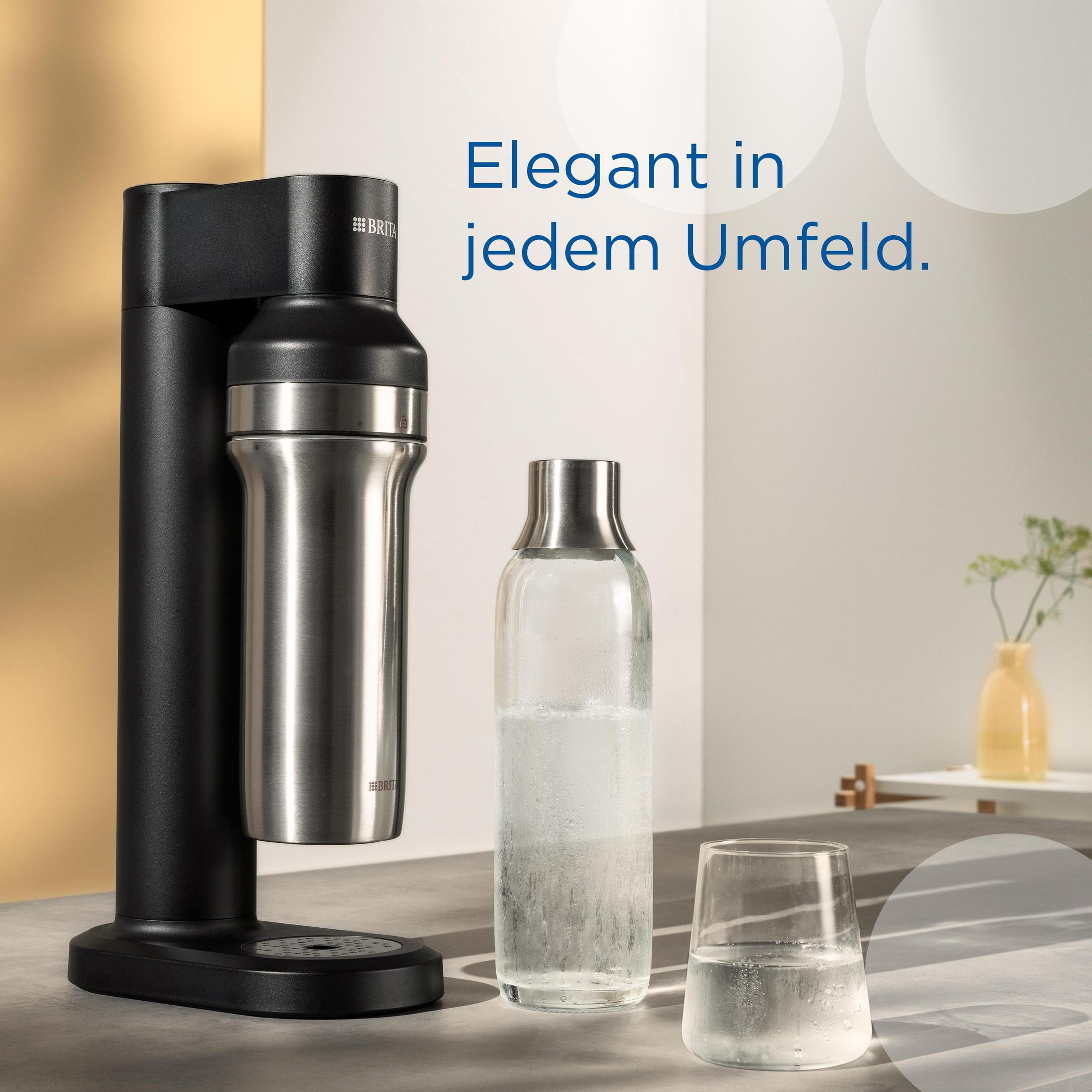 Eine schwarze Wassermaschine mit einer silbernen Flasche und einem Glas Wasser daneben auf hellem Hintergrund.