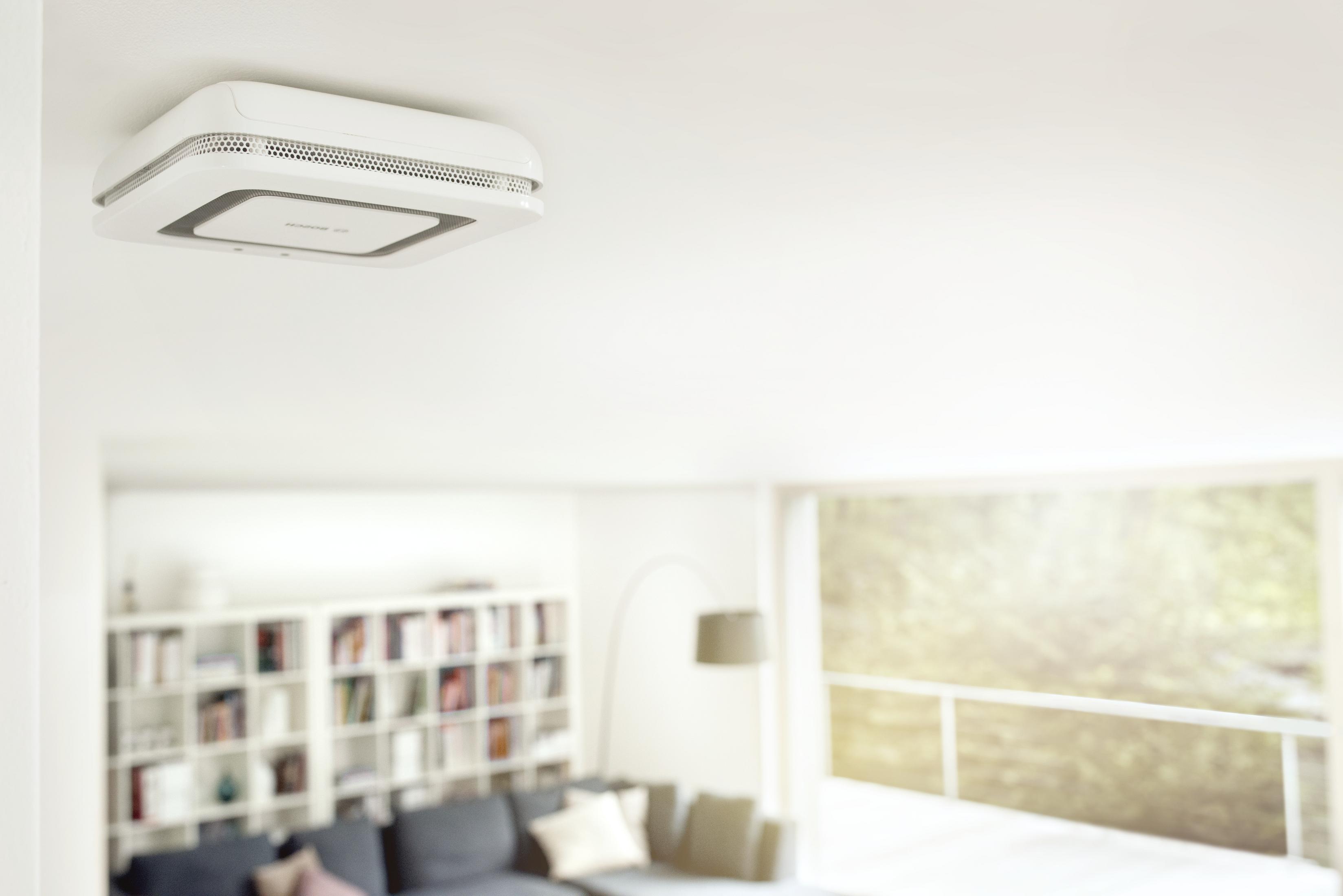 Weißer Rauchmelder an weißer Decke, Wohnzimmer im Hintergrund.