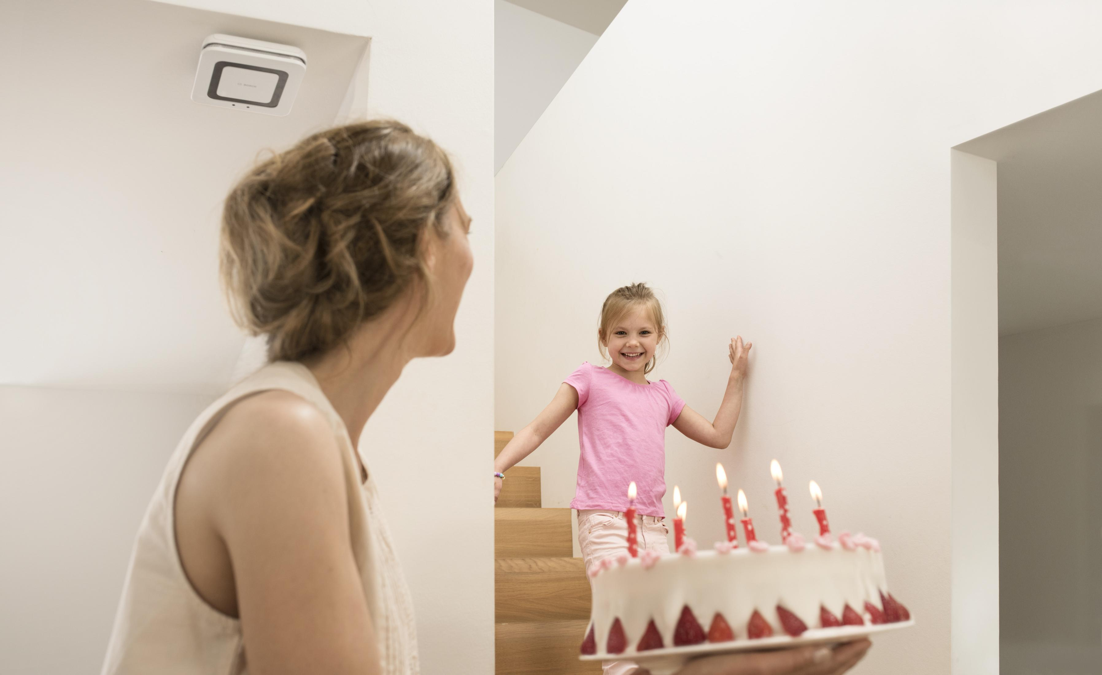 Eine Frau und ein Mädchen halten eine Geburtstagstorte mit Kerzen. Weiße Wände und Treppe im Hintergrund.