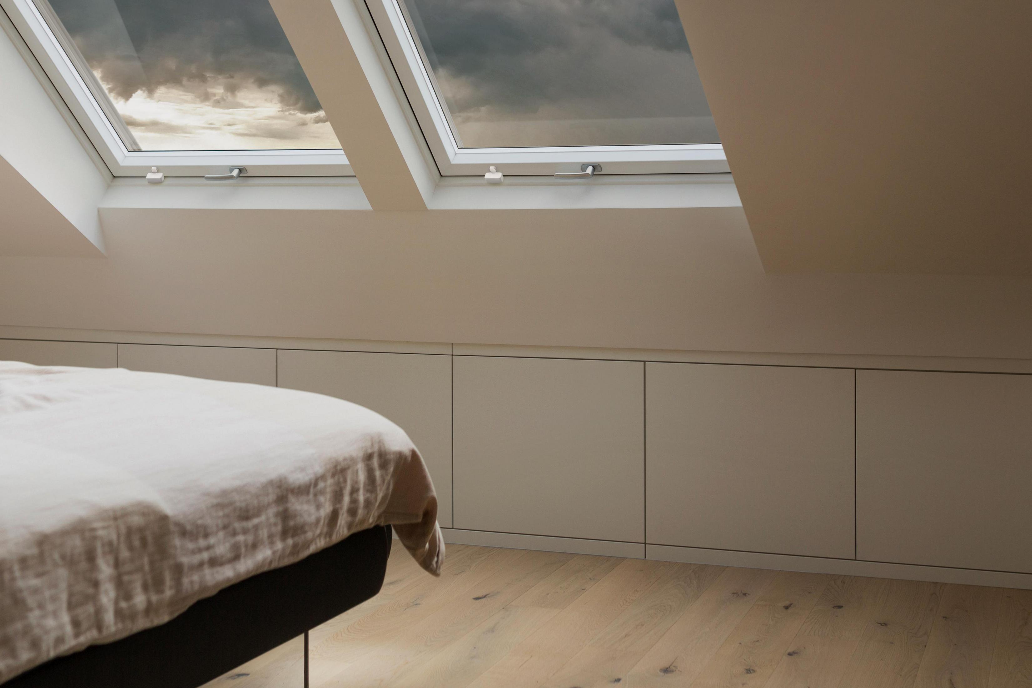 Sypialnia z łóżkiem, białymi ścianami, oknami dachowymi i drewnianą podłogą. Widoczne pochmurne niebo.