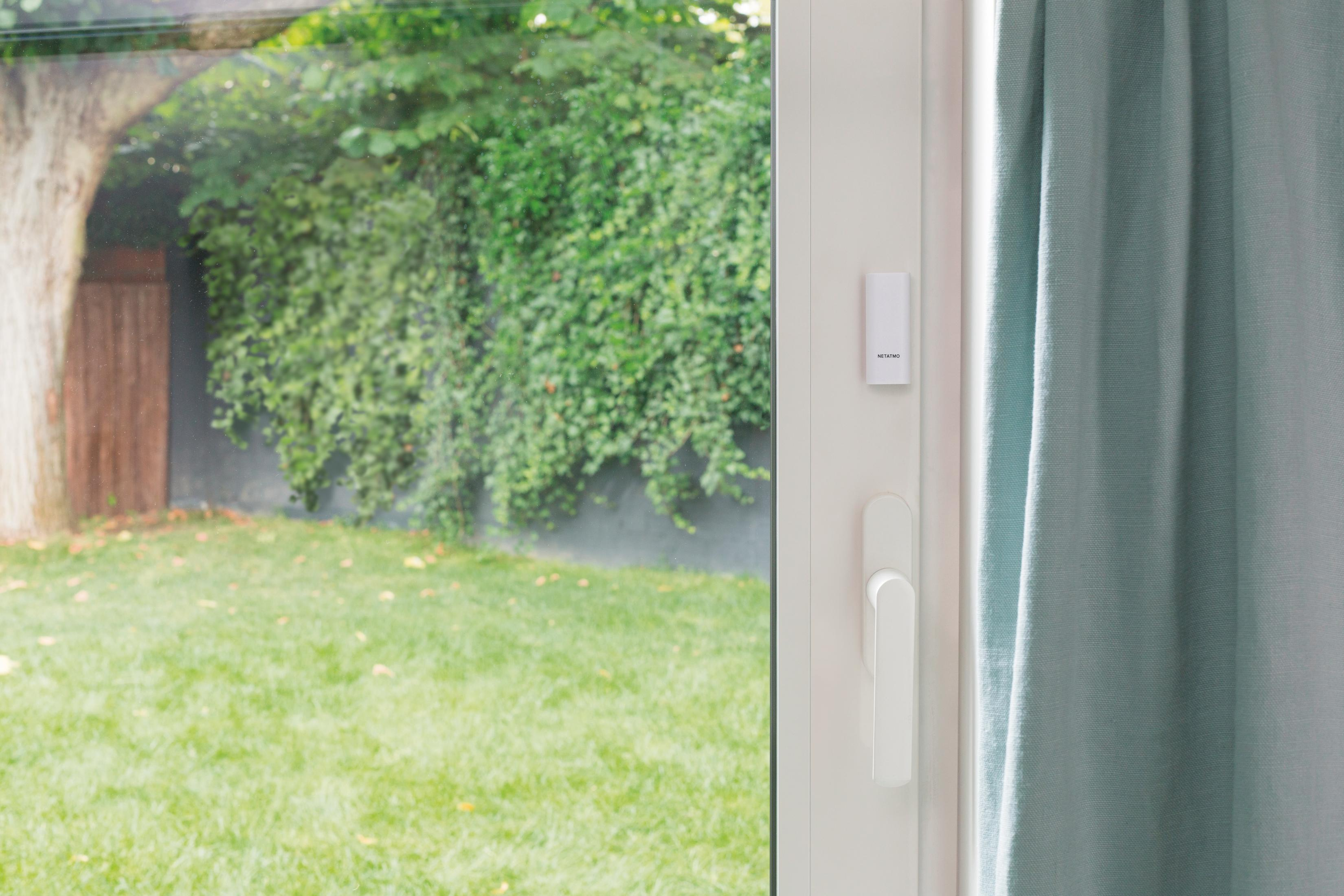 Maniglia finestra con sensore bianco e tenda accanto, vista giardino verde.