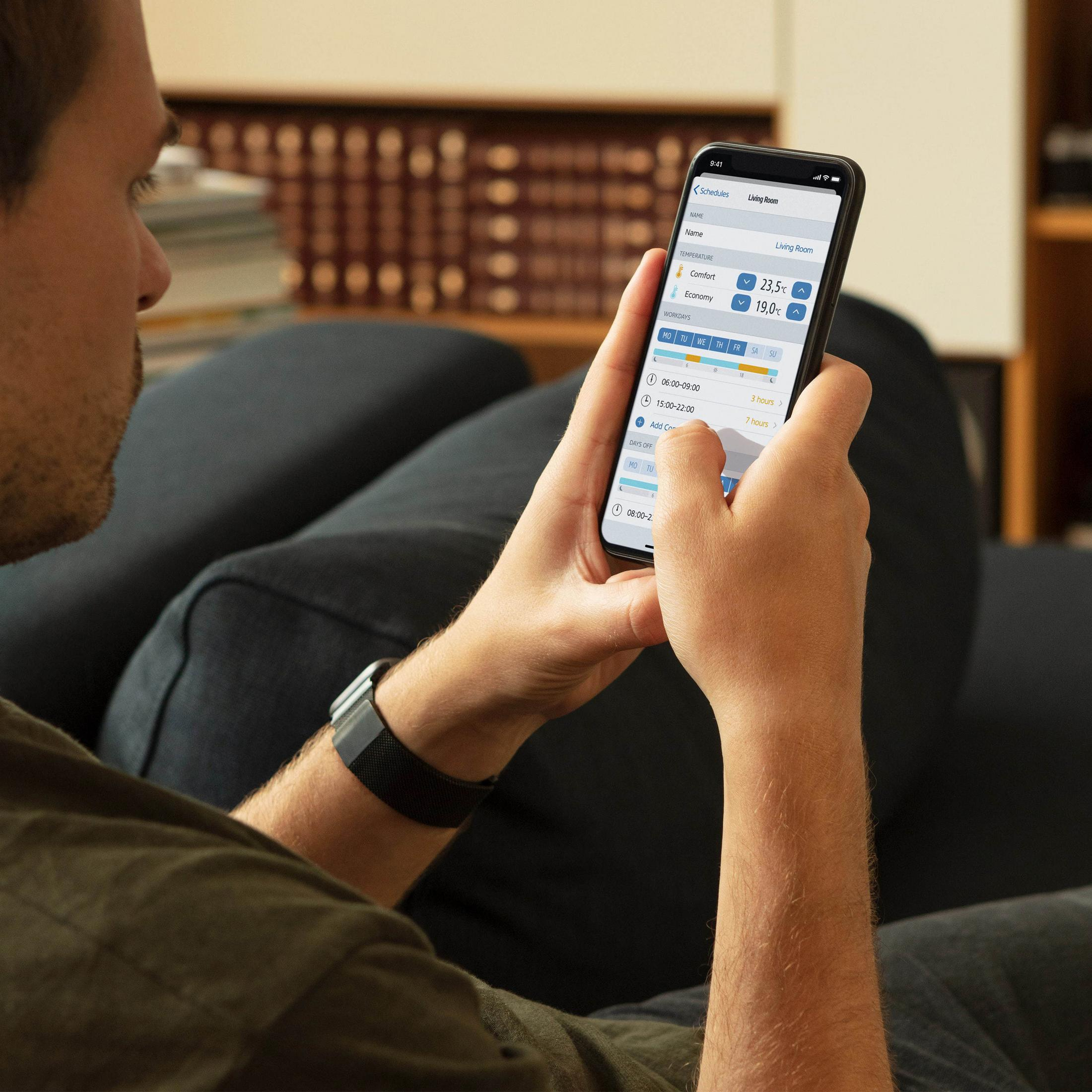 Mann benutzt Smartphone, App-Oberfläche sichtbar. Auf dunklem Sofa sitzend.