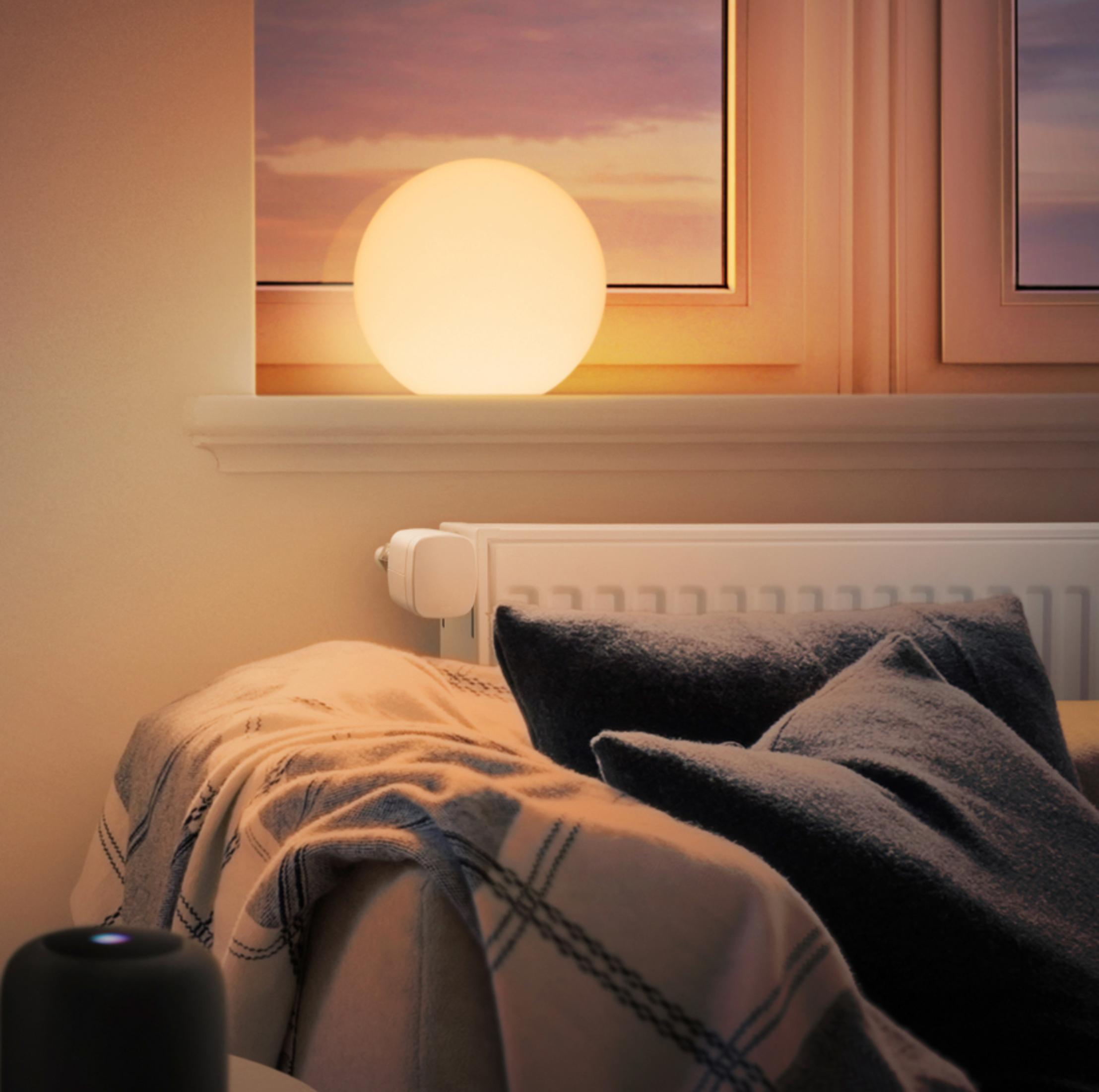 Eine runde, leuchtende Lampe auf einer Fensterbank. Kissen und eine Decke auf einem Bett. Warme Beleuchtung.