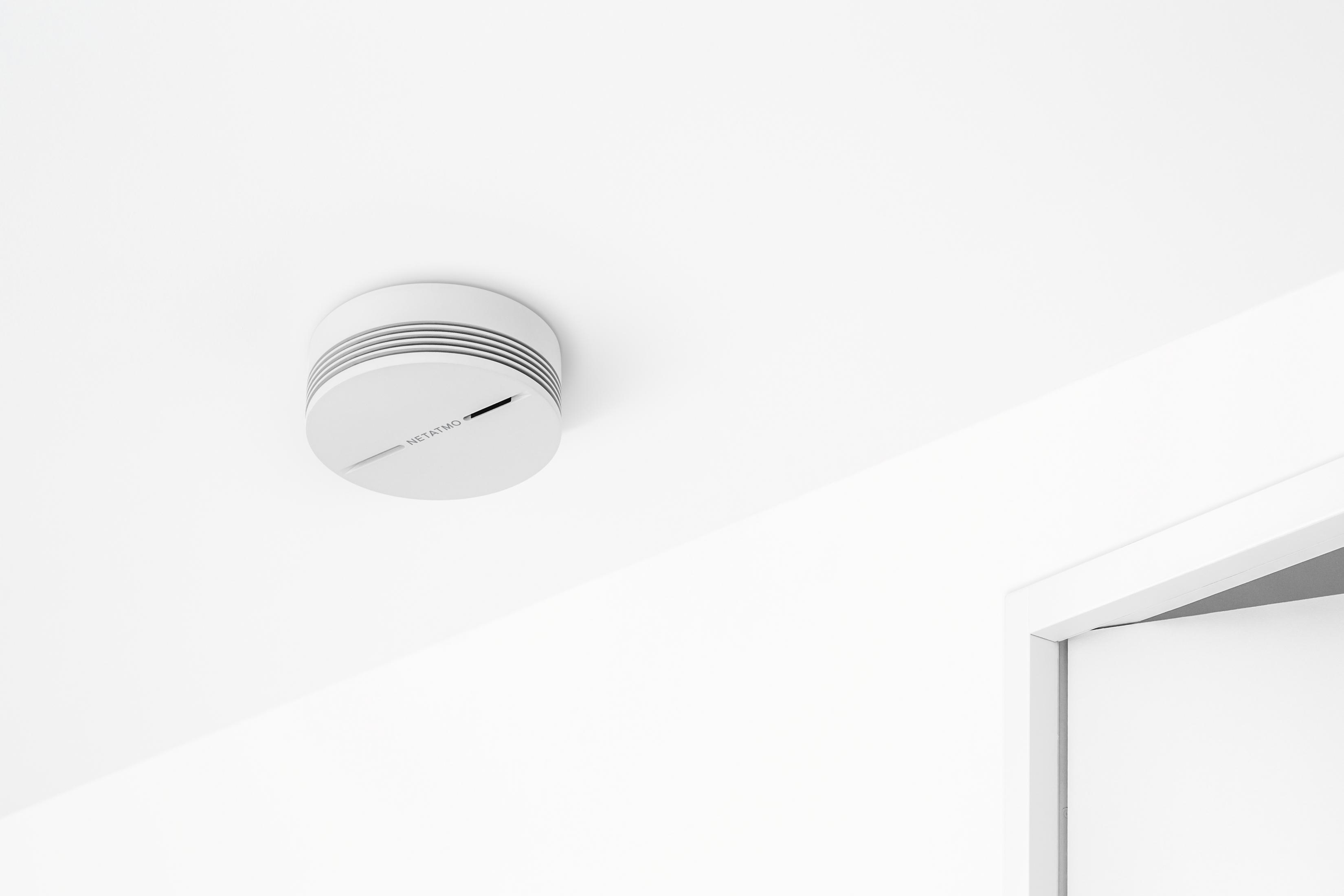 Biały czujnik dymu Netatmo zamontowany na białym suficie, z białą ościeżnicą.