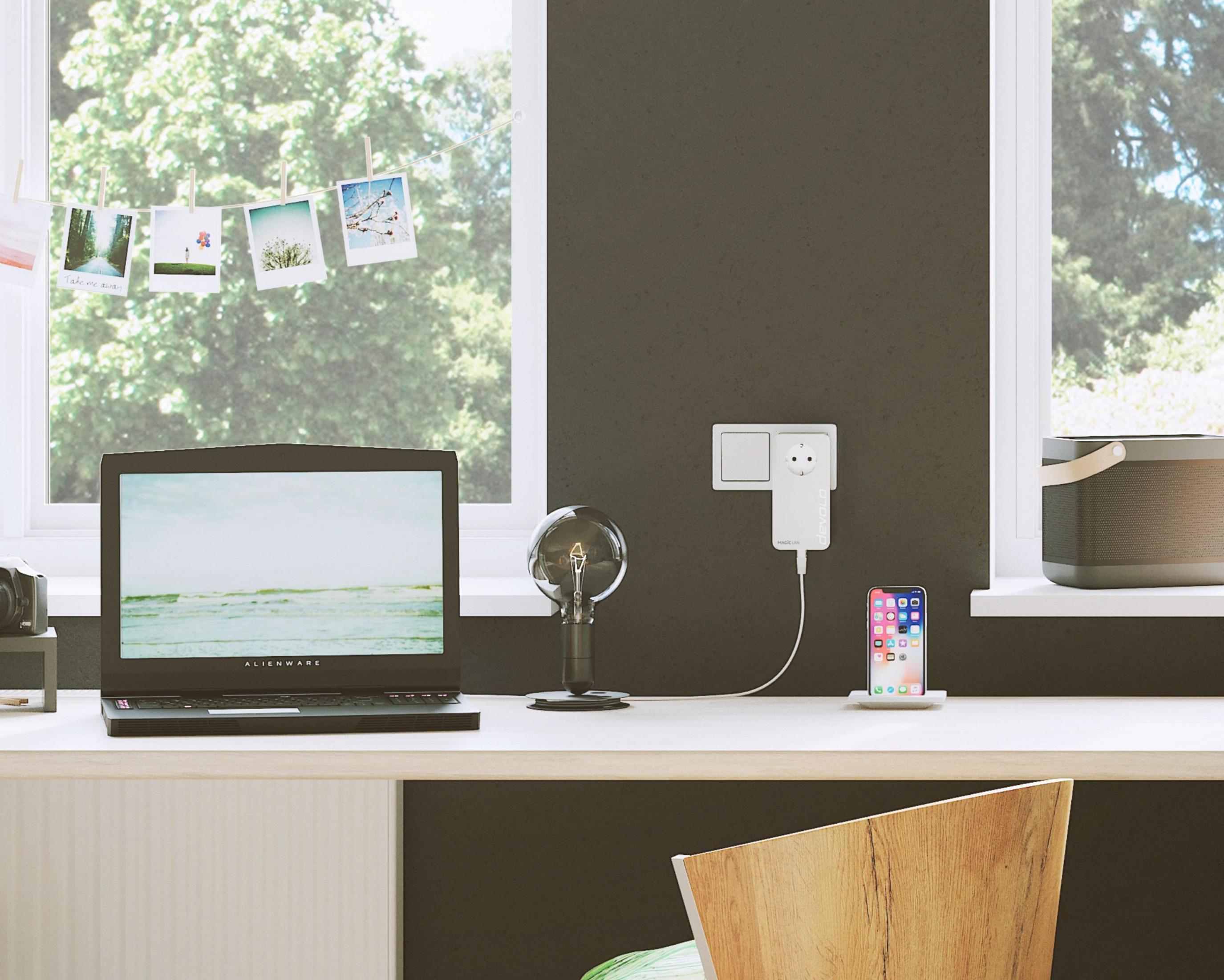 Biurko z laptopem, telefonem, lampą i oknem. Telefon się ładuje. Widok na zewnątrz przez okno.