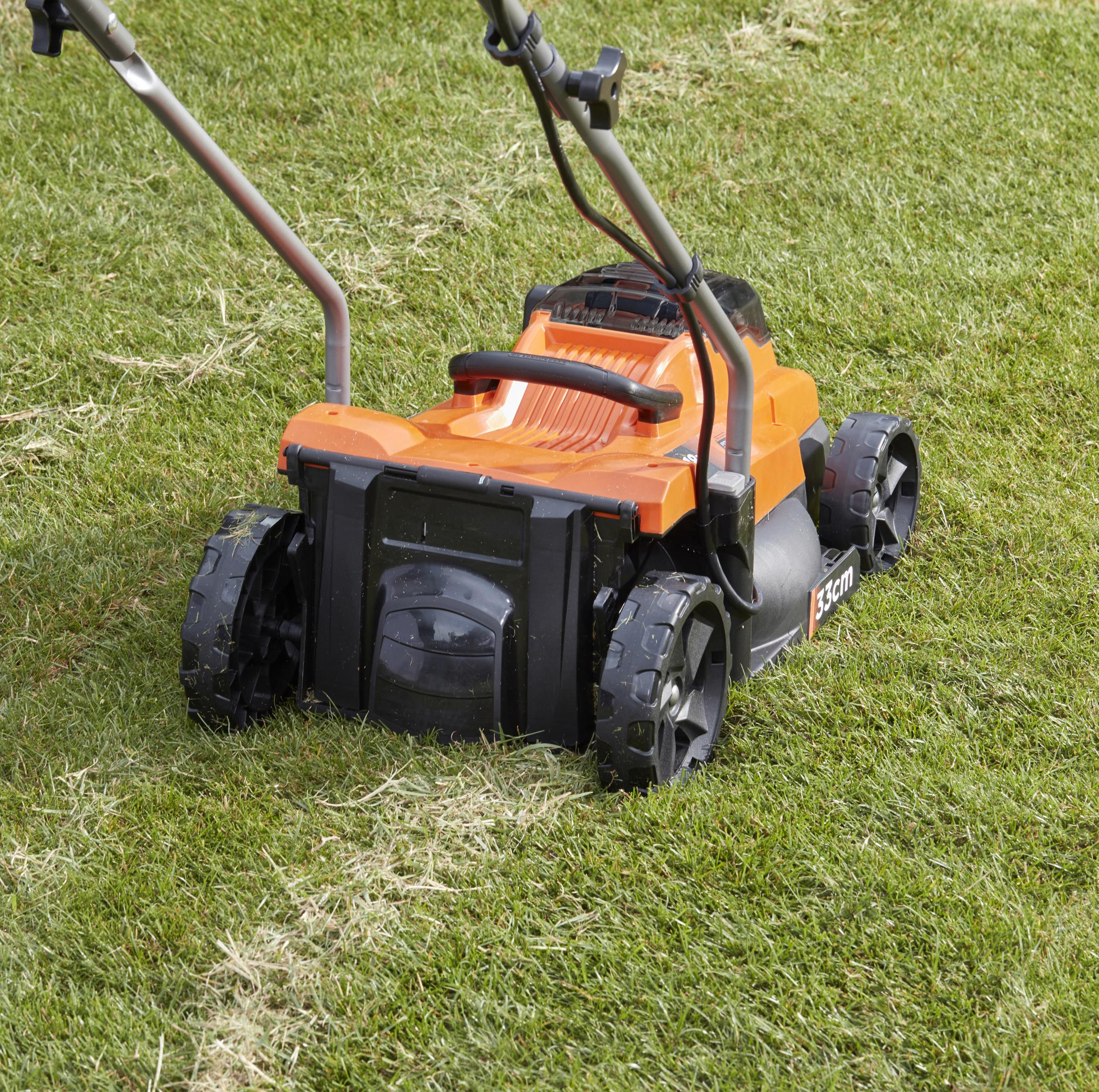Ein orangefarbener und schwarzer Rasenmäher schneidet grünes Gras. Der Mäher hat eine 33cm Markierung.