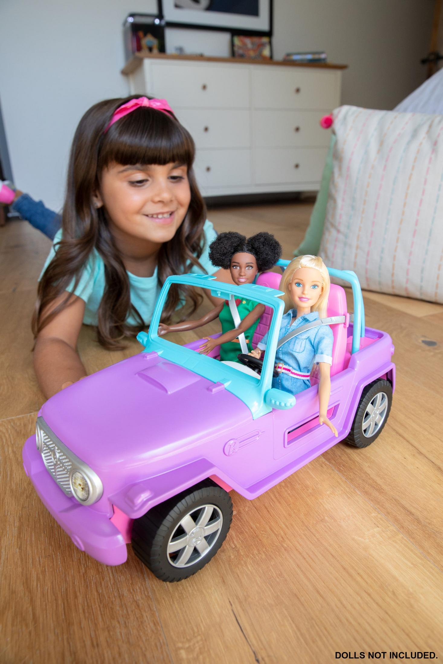 Niña con diadema rosa mira un coche de juguete morado con muñecas. Muñecas no incluidas.