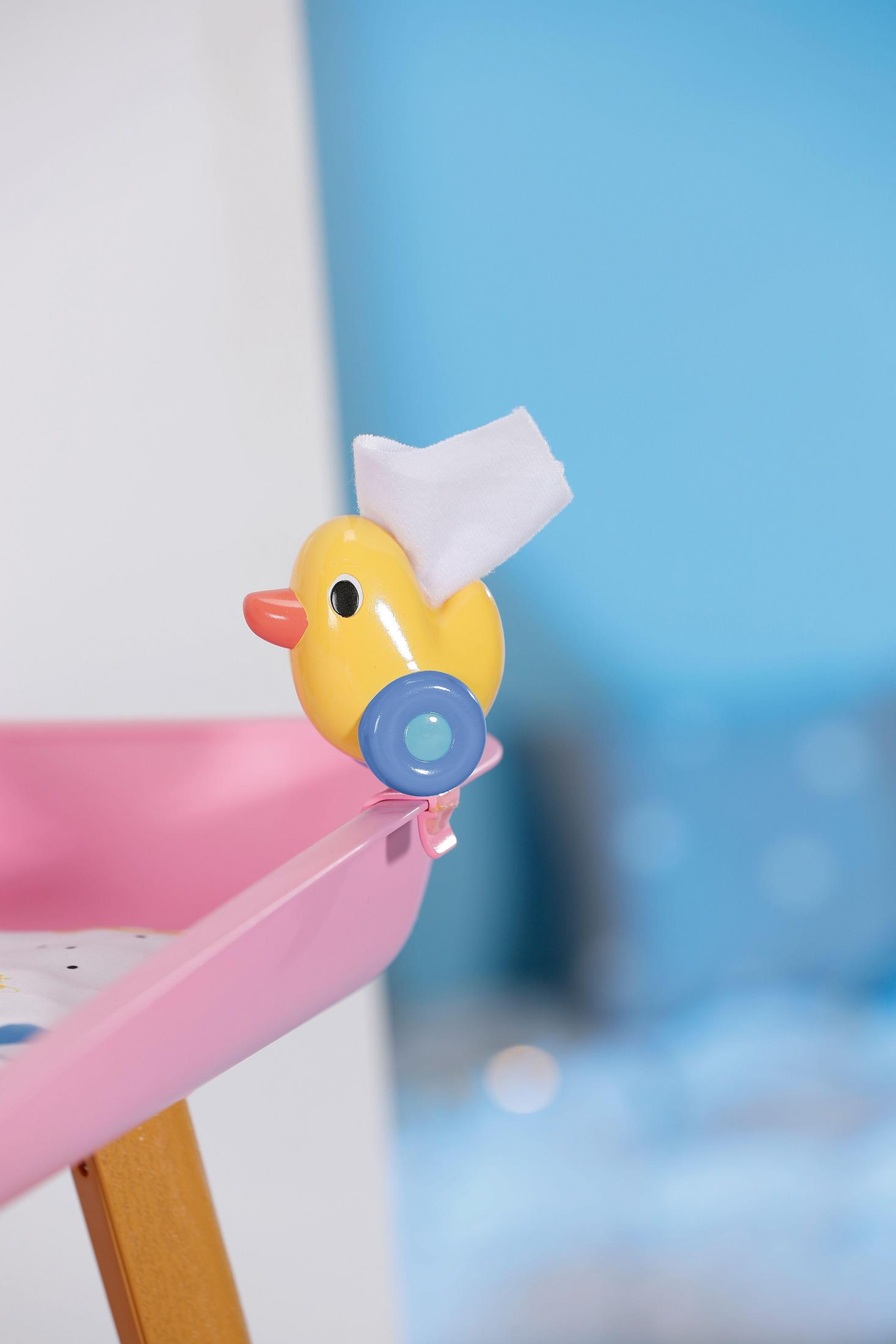 Juguete de pato amarillo con un pañuelo blanco sobre una mesa cambiador rosa. Fondo de pared azul.