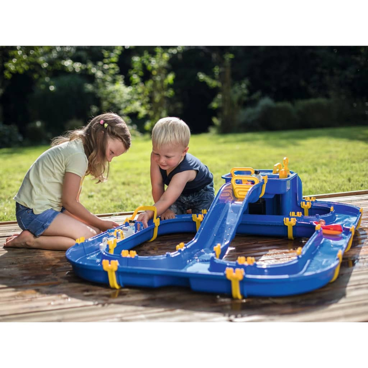 Dwoje dzieci bawiących się zestawem do zabawy w wodzie na drewnianym tarasie na zewnątrz. Z tyłu zielona trawa.